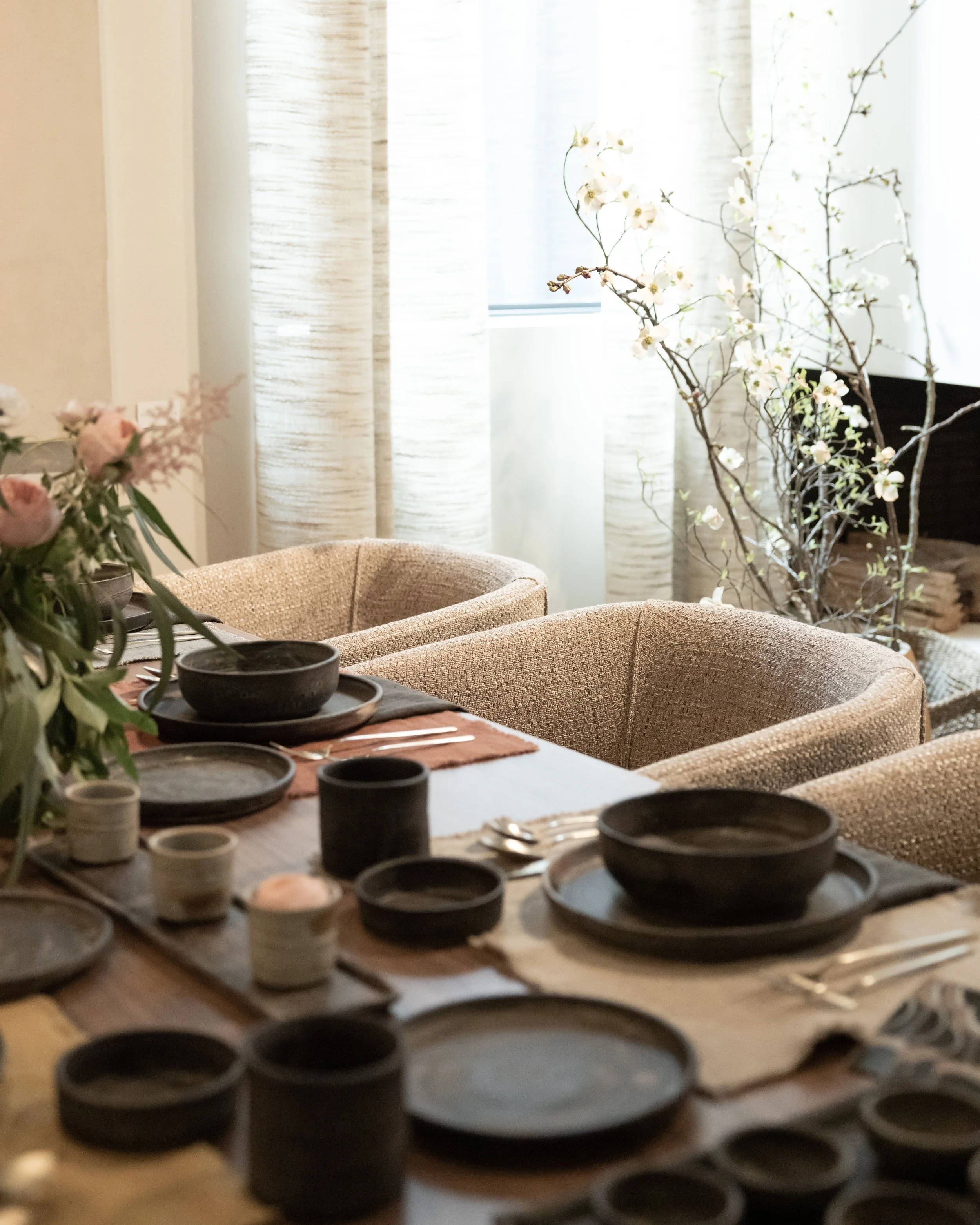 A dining room with a table set with black and white dishes, surrounded by beige upholstered chairs. In the background are light-colored curtains, a window, and a tall branch with white blossoms.