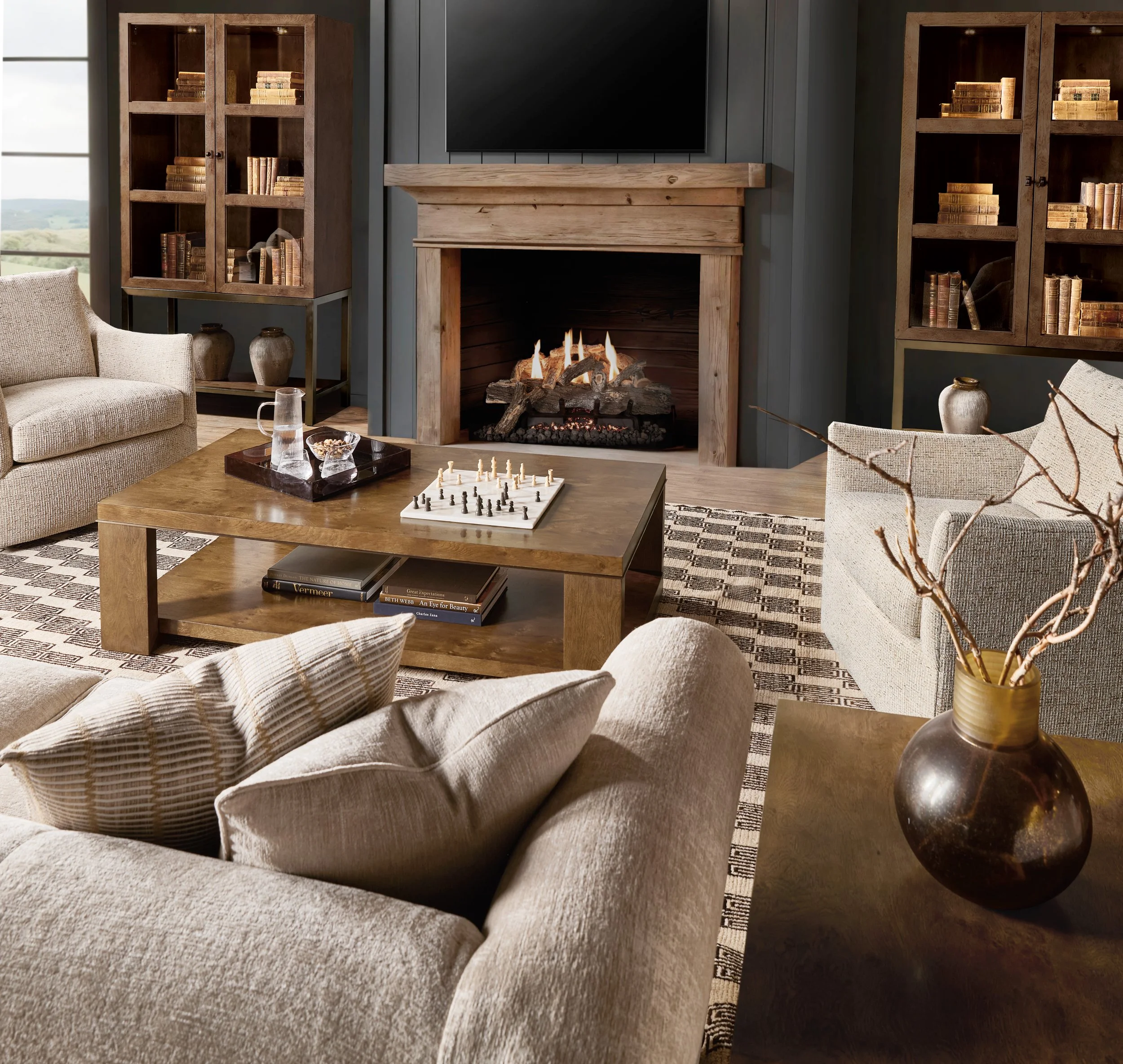 Living room with a beige sectional sofa, circular ottoman, fireplace, abstract artwork, hanging light fixture, and small table with a flower arrangement.
