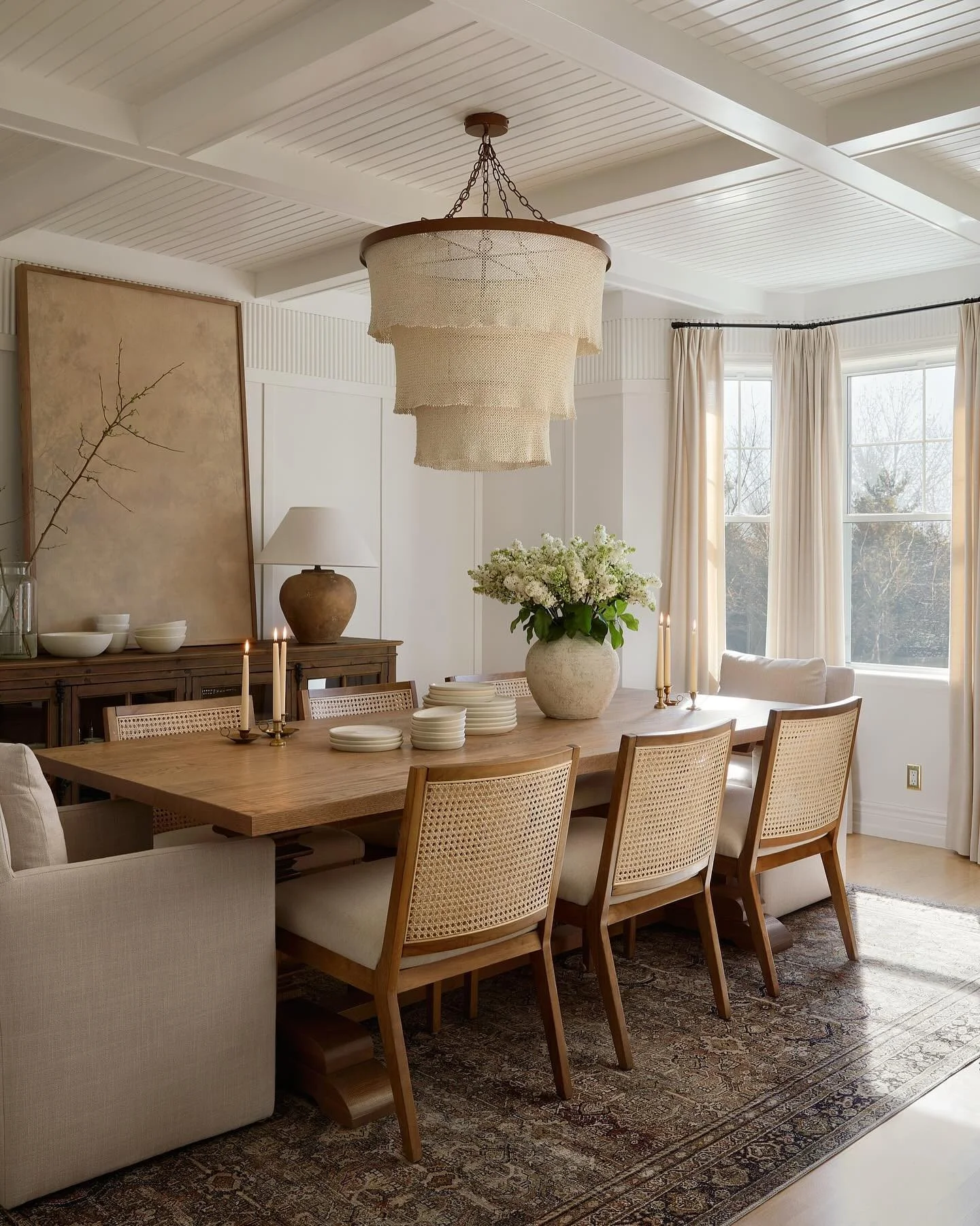 Bright dining room with a large wooden table, cream chairs, a floral centerpiece, candles, a sideboard with bowls, a large painting, and large windows with cream curtains.