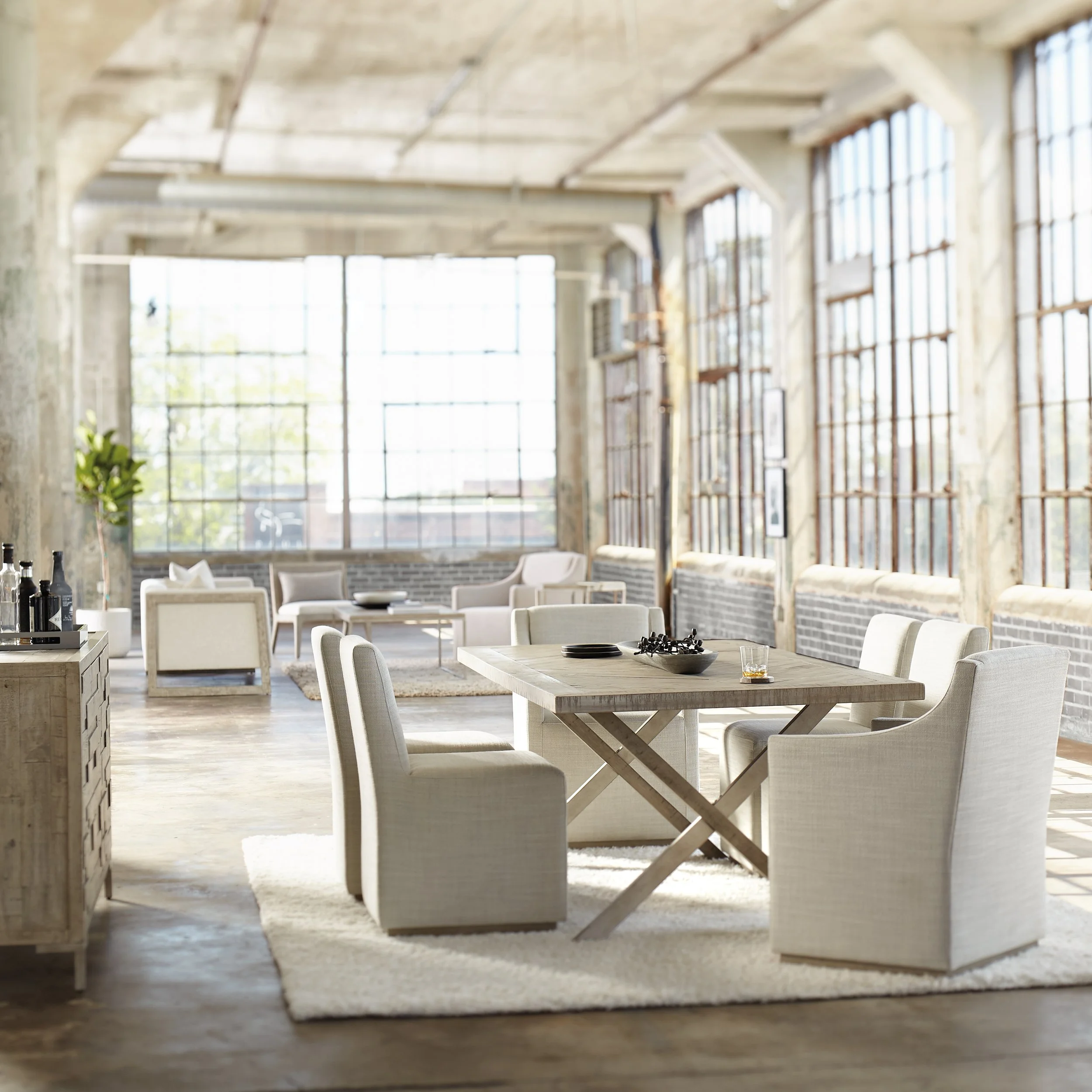 Brightly lit industrial-style interior with large windows, high ceilings, and minimalist furniture including tables and chairs, with a rugged, unfinished aesthetic.