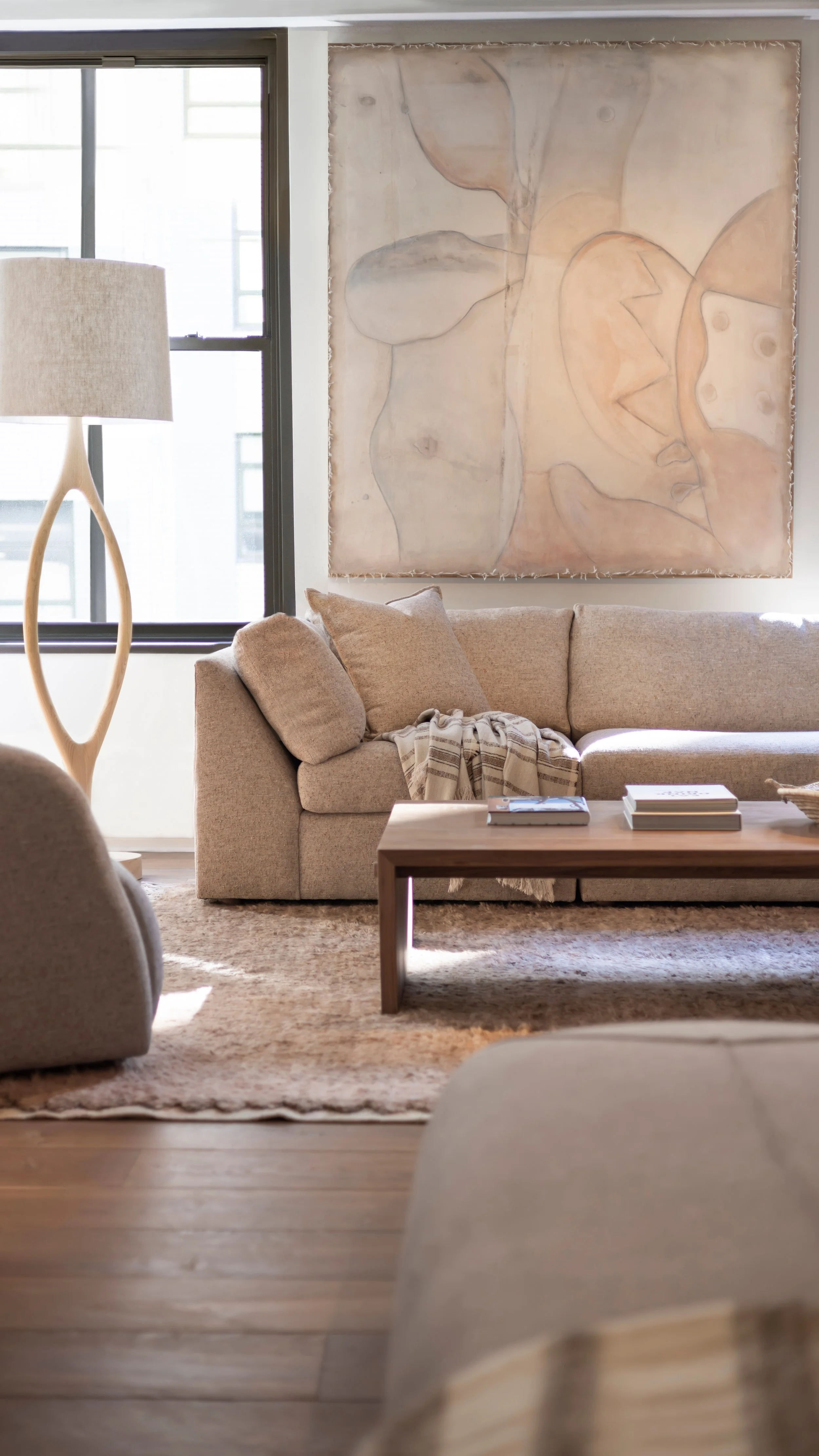 Living room with beige sofa, wooden coffee table, large abstract face artwork on wall, beige lamp, and natural light coming through a window.