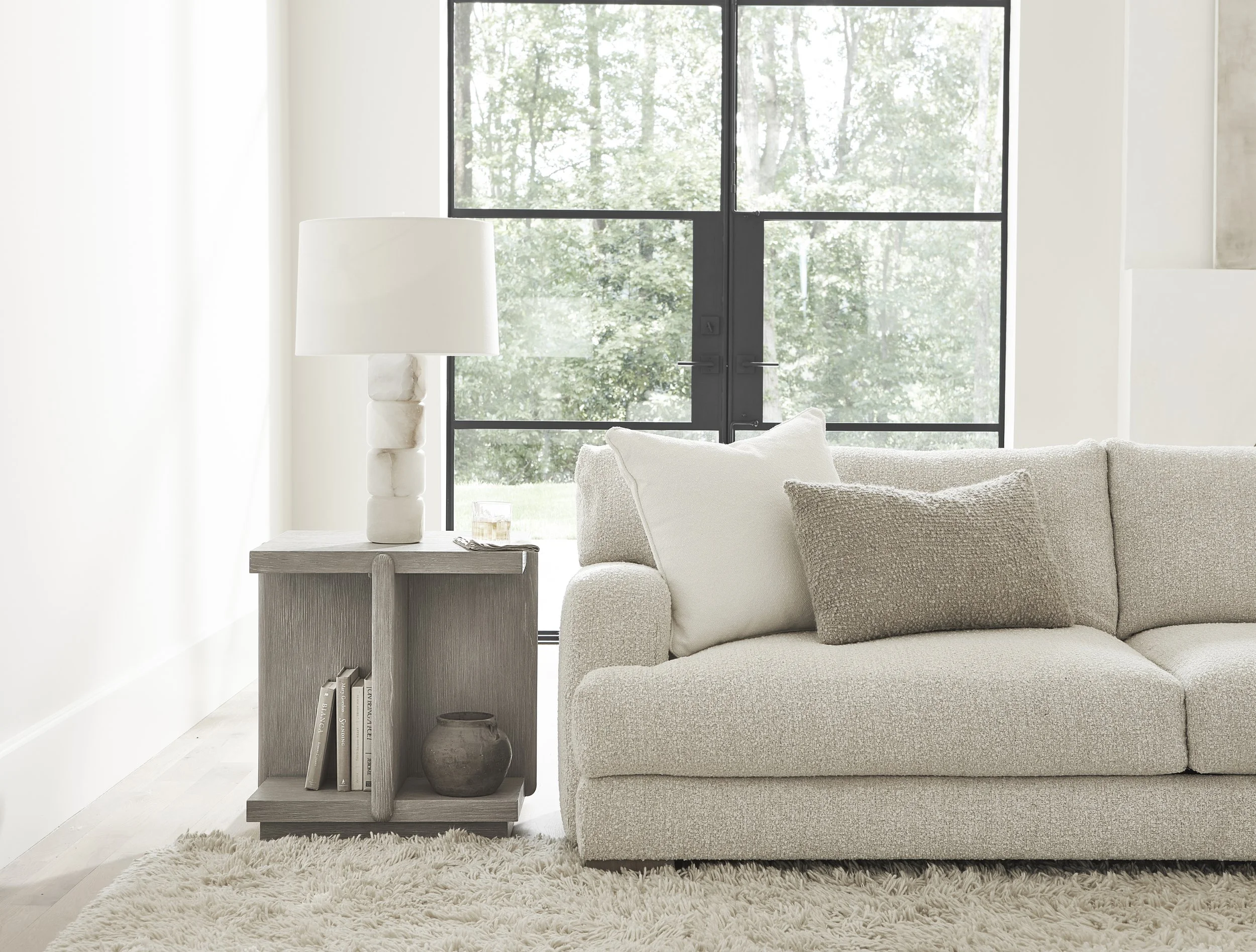 A modern living room with a beige textured sofa, a wooden side table with books and a decorative vase, a white table lamp, large black framed windows showing green trees outside, a white shag rug, and white walls.