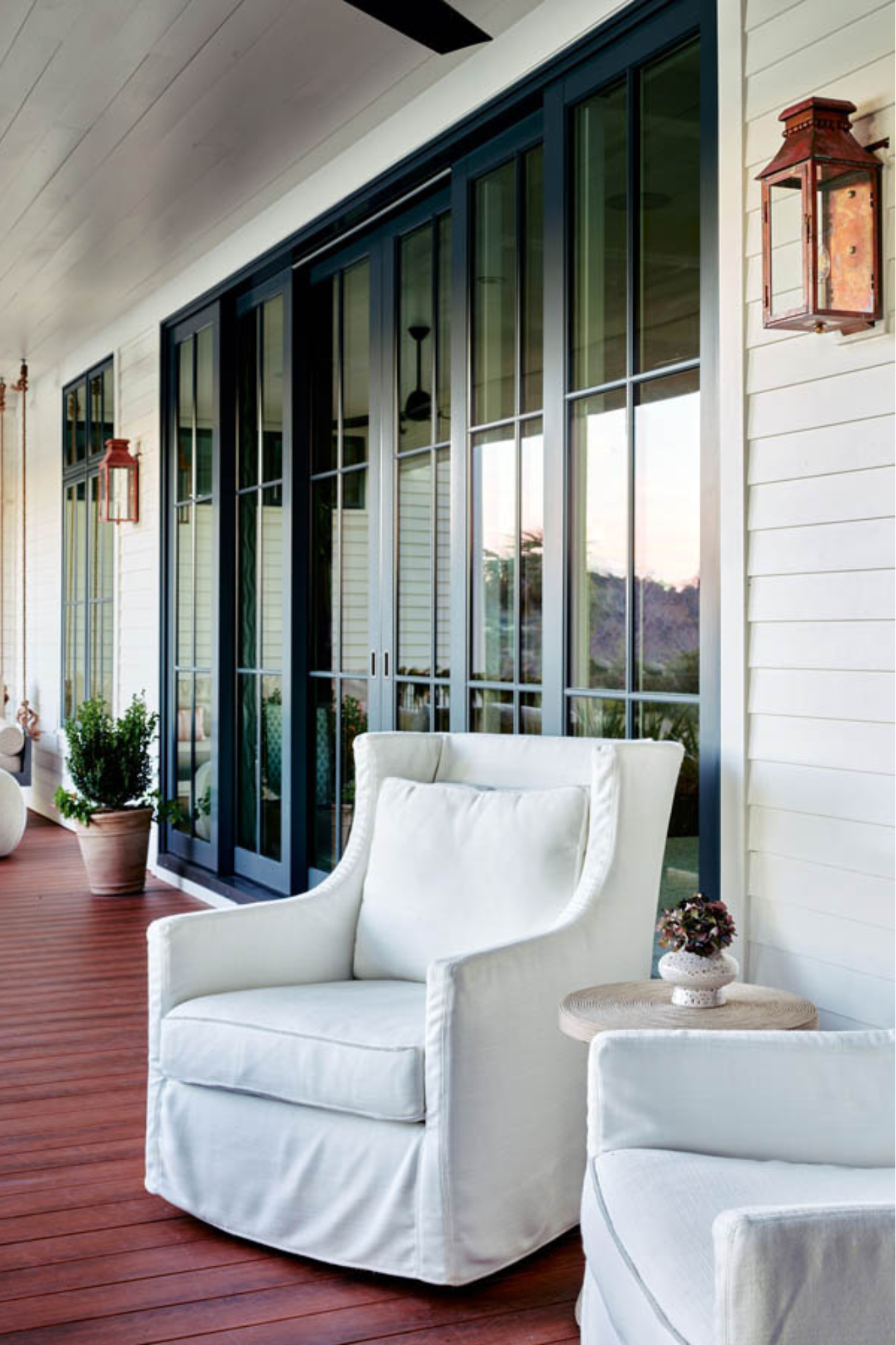 A porch with white upholstered chairs, a small round side table with a flower pot, potted plants, and a large glass sliding door with black frames leading to the interior of a house.