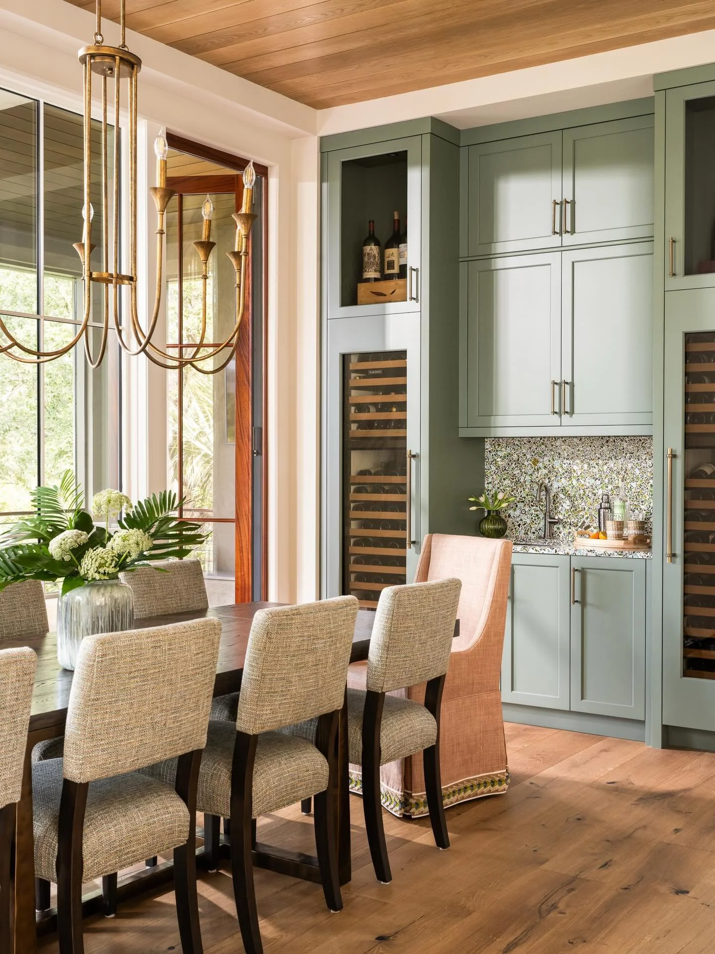 A dining room with a wooden table surrounded by beige upholstered chairs. A large glass vase with white flowers and green leaves sits on the table. To the side, there is a built-in green cabinet with wine bottles and a wine fridge. A window with black metal framing allows natural light to fill the room, and a gold chandelier hangs from the wooden ceiling.