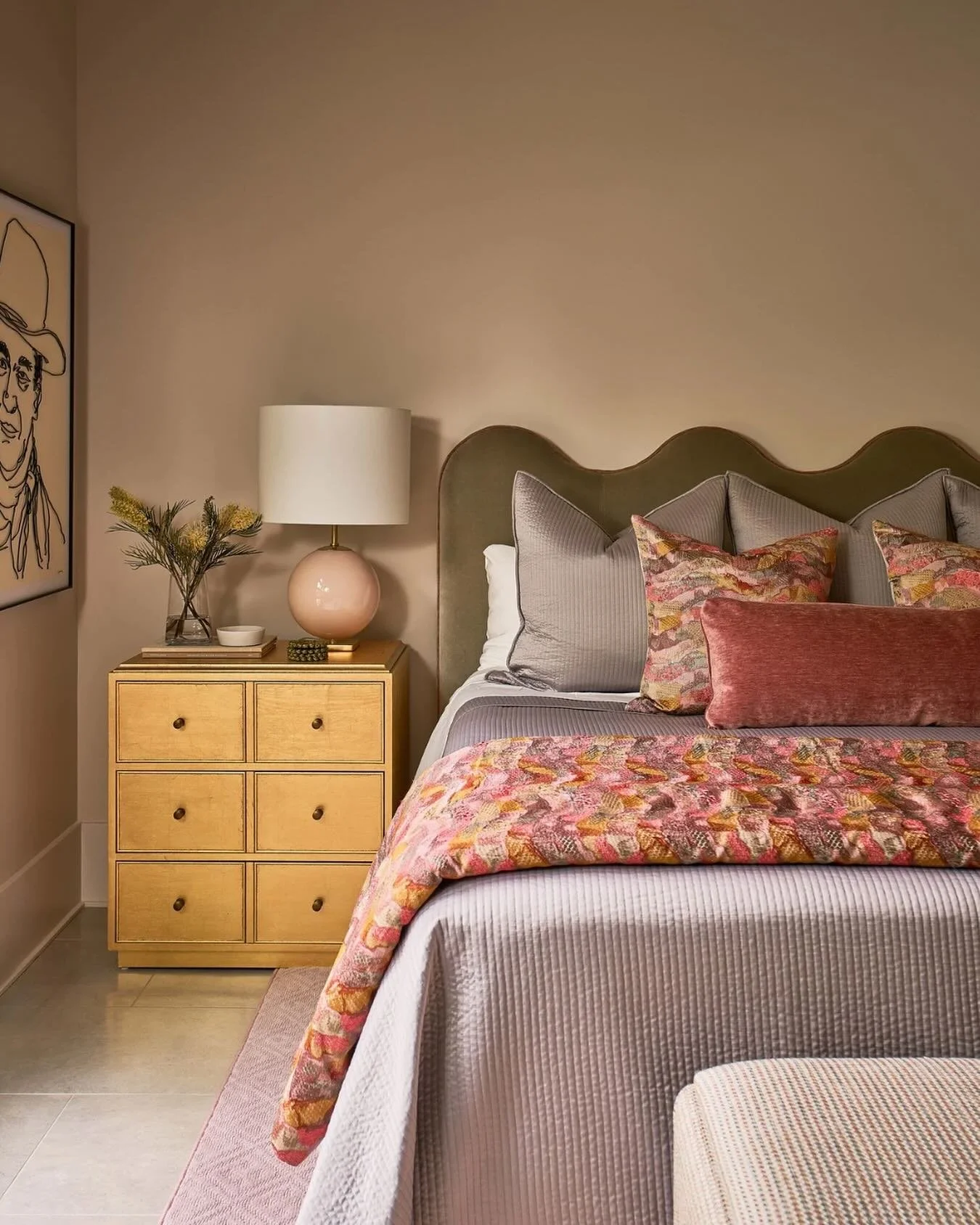 A cozy living room with pink and orange striped walls and ceiling, a white sofa with colorful pillows, a floral rug, a coffee table with books and a vase of flowers, and large windows allowing natural light.