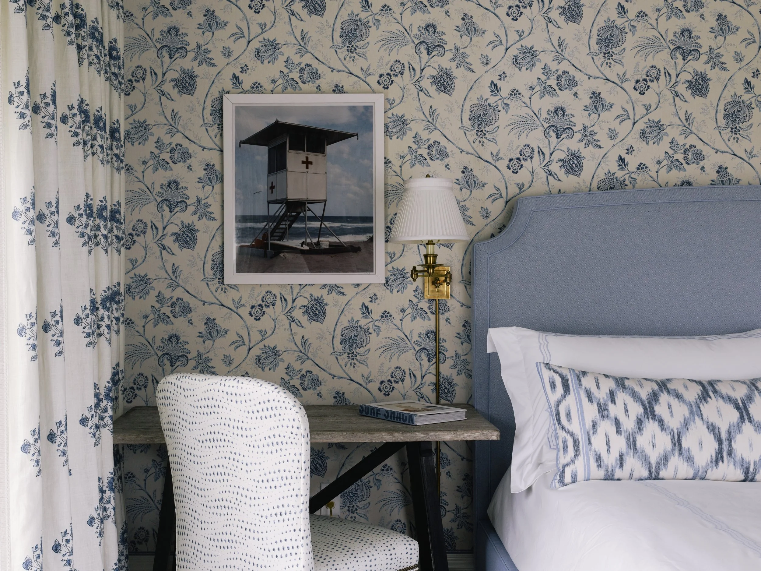 A bedroom with floral blue and white wallpaper and matching curtains. There is a framed picture of a lifeguard station on the wall, a brass wall lamp, a bed with a blue headboard and white and blue striped bedding, a polka dot chair, a wooden desk, and a magazine titled "Surf Shaun."