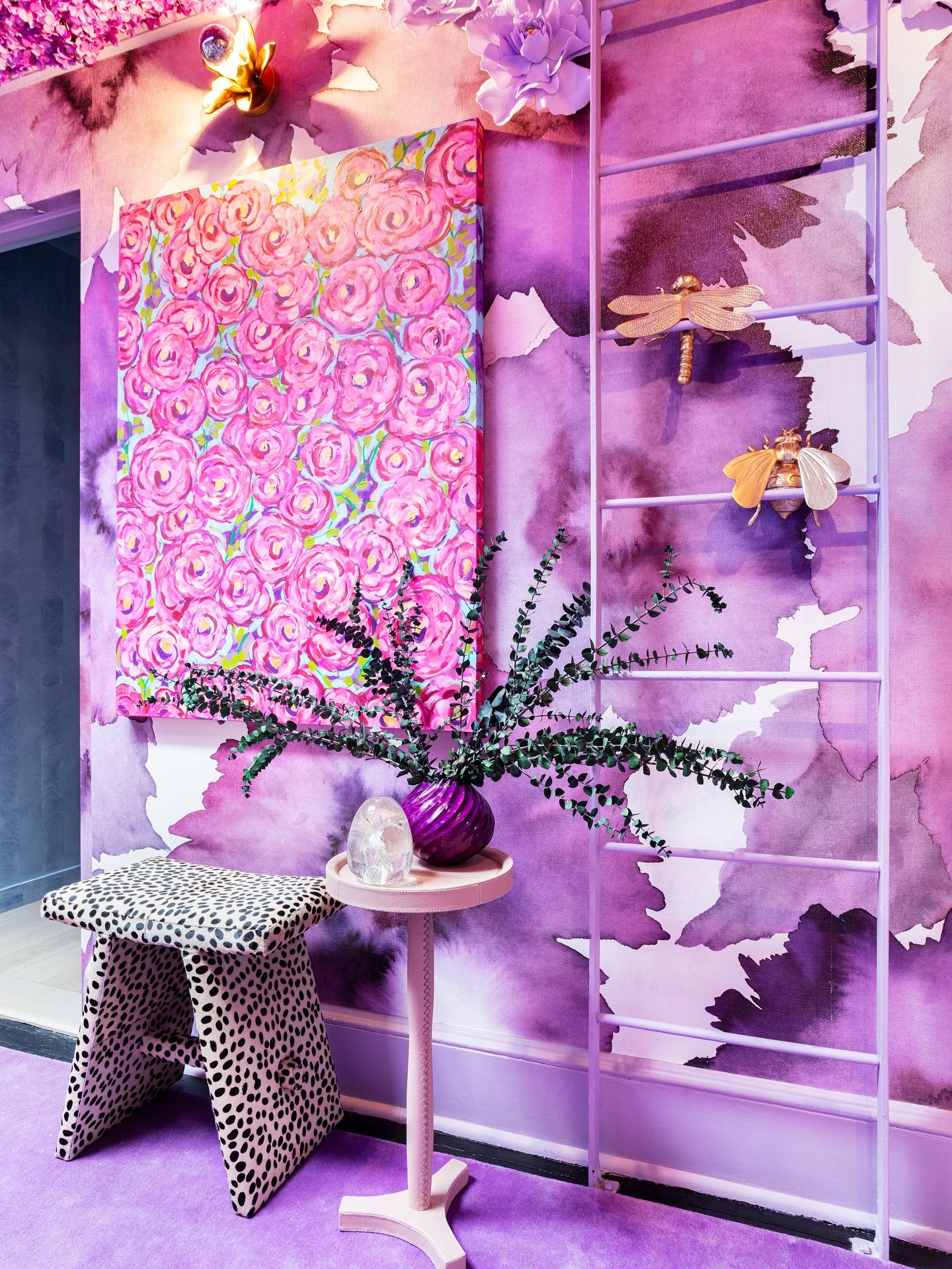 Interior decor with a purple floral theme, featuring a pink canvas with pink roses, a purple vase with green foliage, a small pink table, a polka-dotted stool, and a purple ladder with dragonfly and bee ornaments, all set against a purple watercolor-style wall.