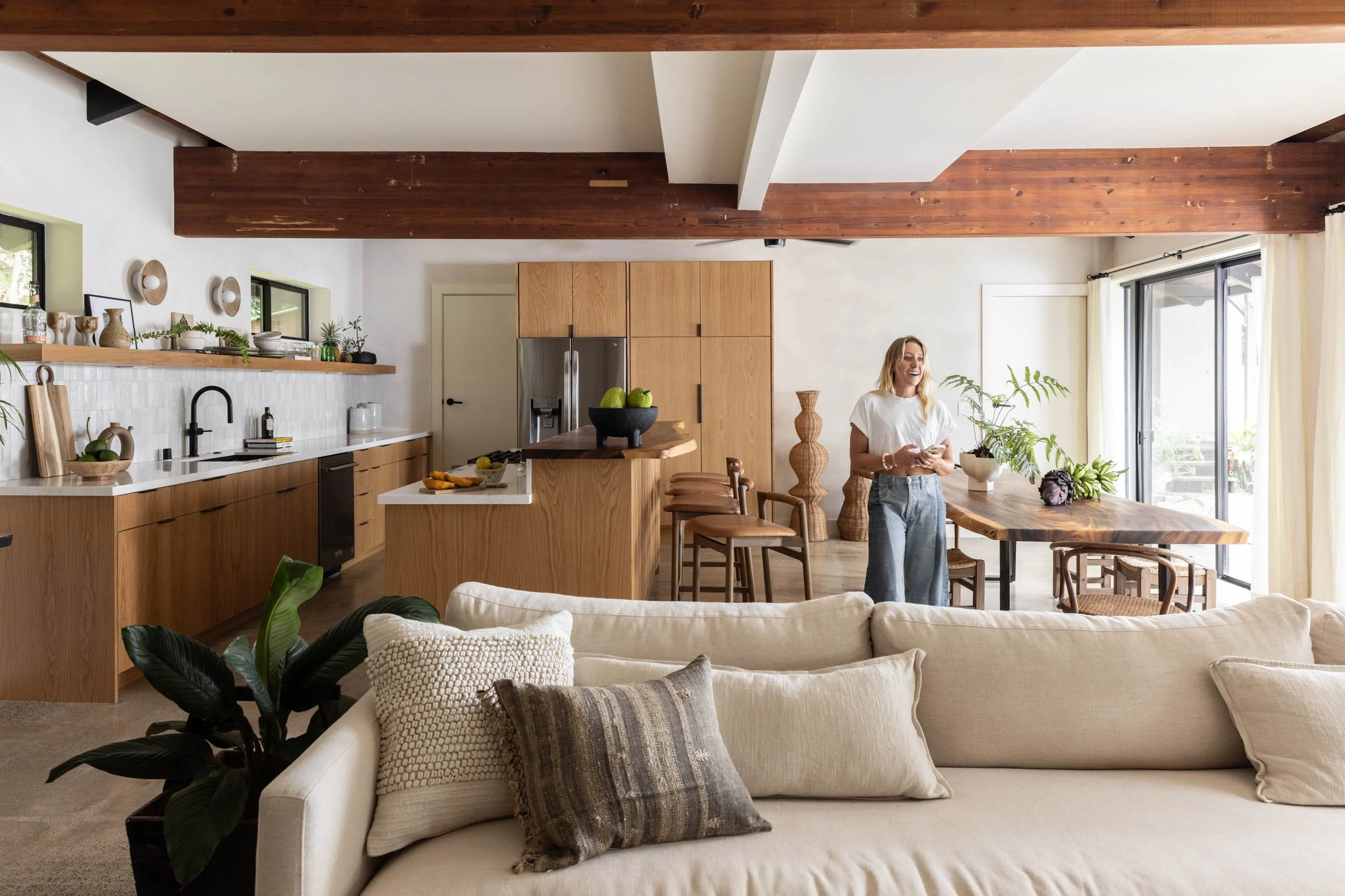 Open-concept kitchen and living area with a woman smiling, standing near a dining table. The kitchen has wooden cabinets, a stainless steel refrigerator, and a white countertop. The living room features a beige sofa with textured and patterned pillows, and a large sliding glass door with curtains letting in natural light.