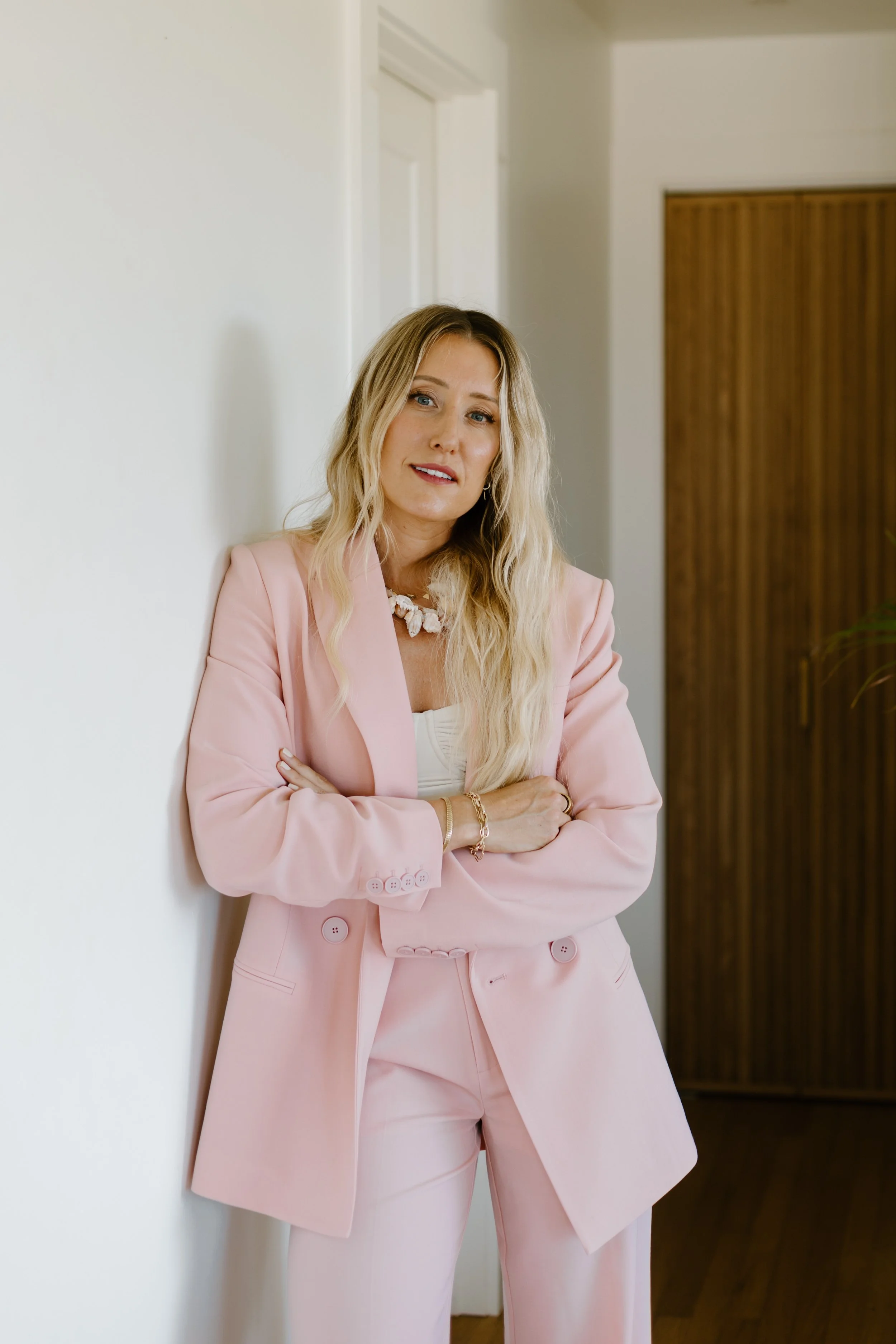 A woman with long blonde hair wearing a pink power suit and a shell necklace, standing in a well lit room