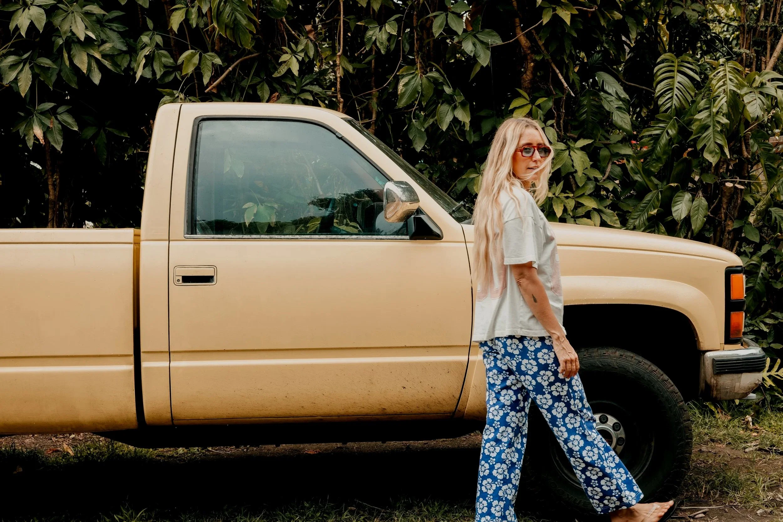 A woman with long blonde hair, wearing sunglasses, a gray T-shirt, and blue floral pants, standing next to a beige pickup truck with the driver's side door closed. She is outdoors with dense green foliage in the background.
