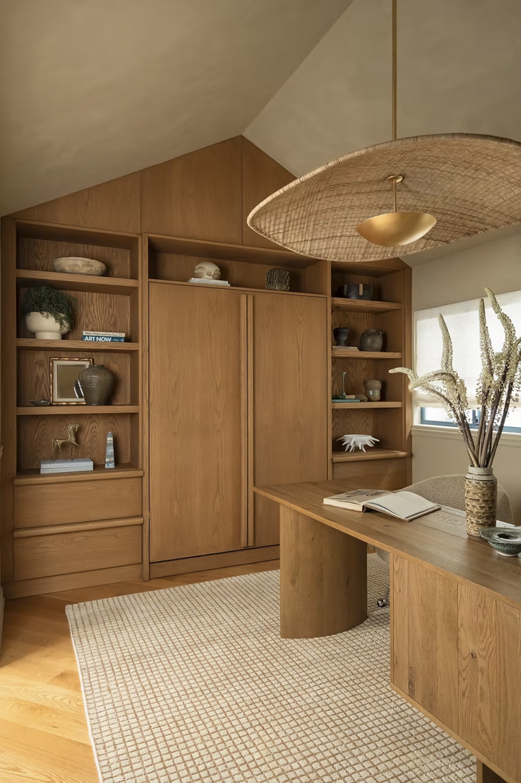 A cozy home office with built-in wooden bookshelves, a wooden desk, a woven ceiling light fixture, and a beige area rug. Decor includes vases, books, and a vase with decorative dried plants.