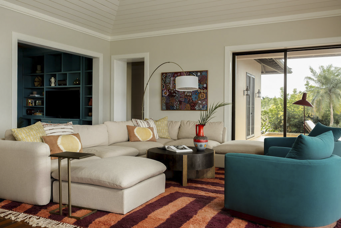 A living room with a white sectional sofa, teal armchair, colorful pillows, a round coffee table with a vase and a book, a large window showing greenery outside, and a built-in blue bookshelf.