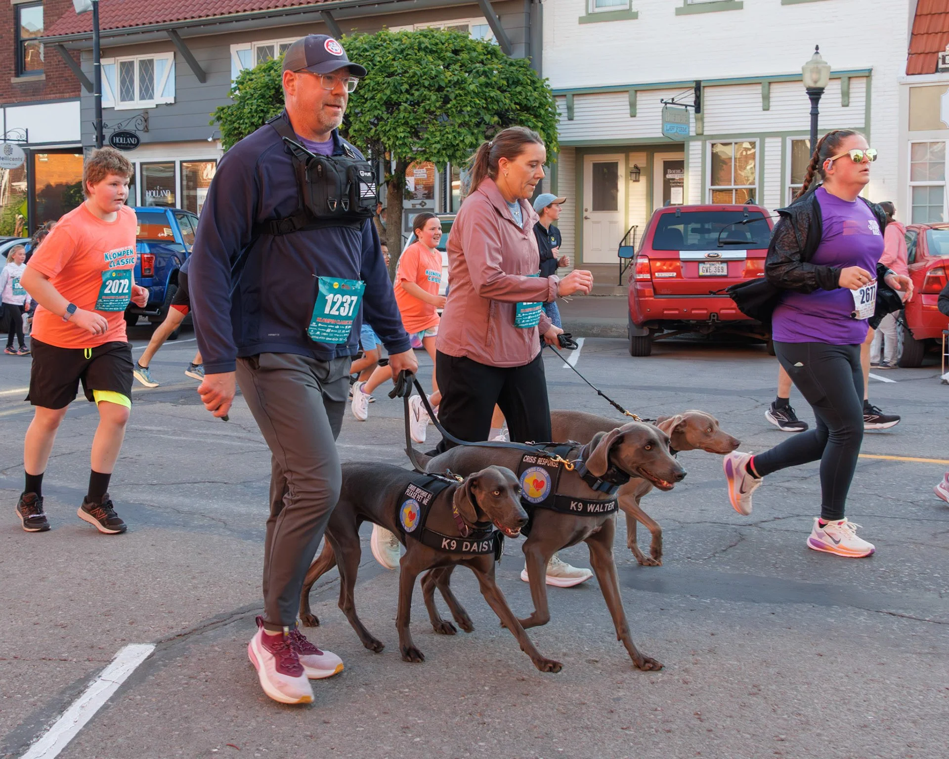A group of people participating in a street event, including a man in a dark jacket and cap, a woman holding a leash with service dogs, children wearing race bibs, and others walking along a city street with storefronts and parked cars in the background.