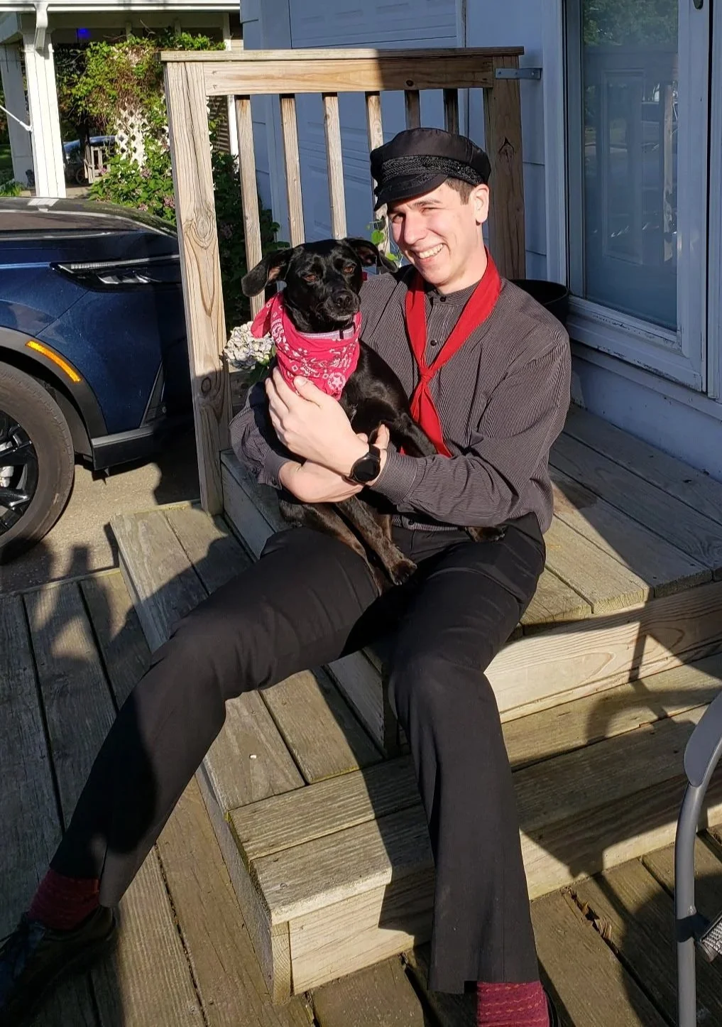 A man sitting on a wooden step holding a black dog wearing a red bandana, smiling in sunlight.