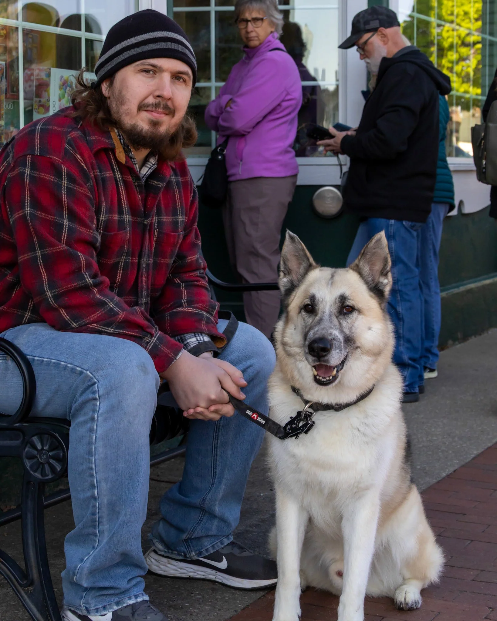 A young man in a red flannel shirt and a beanie sitting on a bench with a happy dog, while two people stand in line behind him, outside a building with large windows.