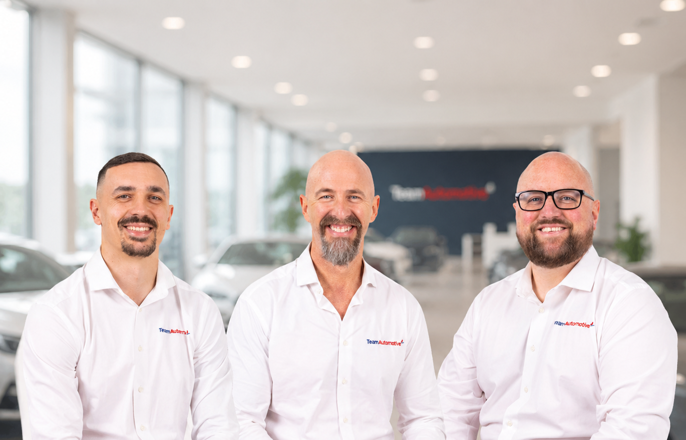 Three men standing in a car dealership showroom, wearing white shirts with "Team Auto" logo, smiling at the camera.
