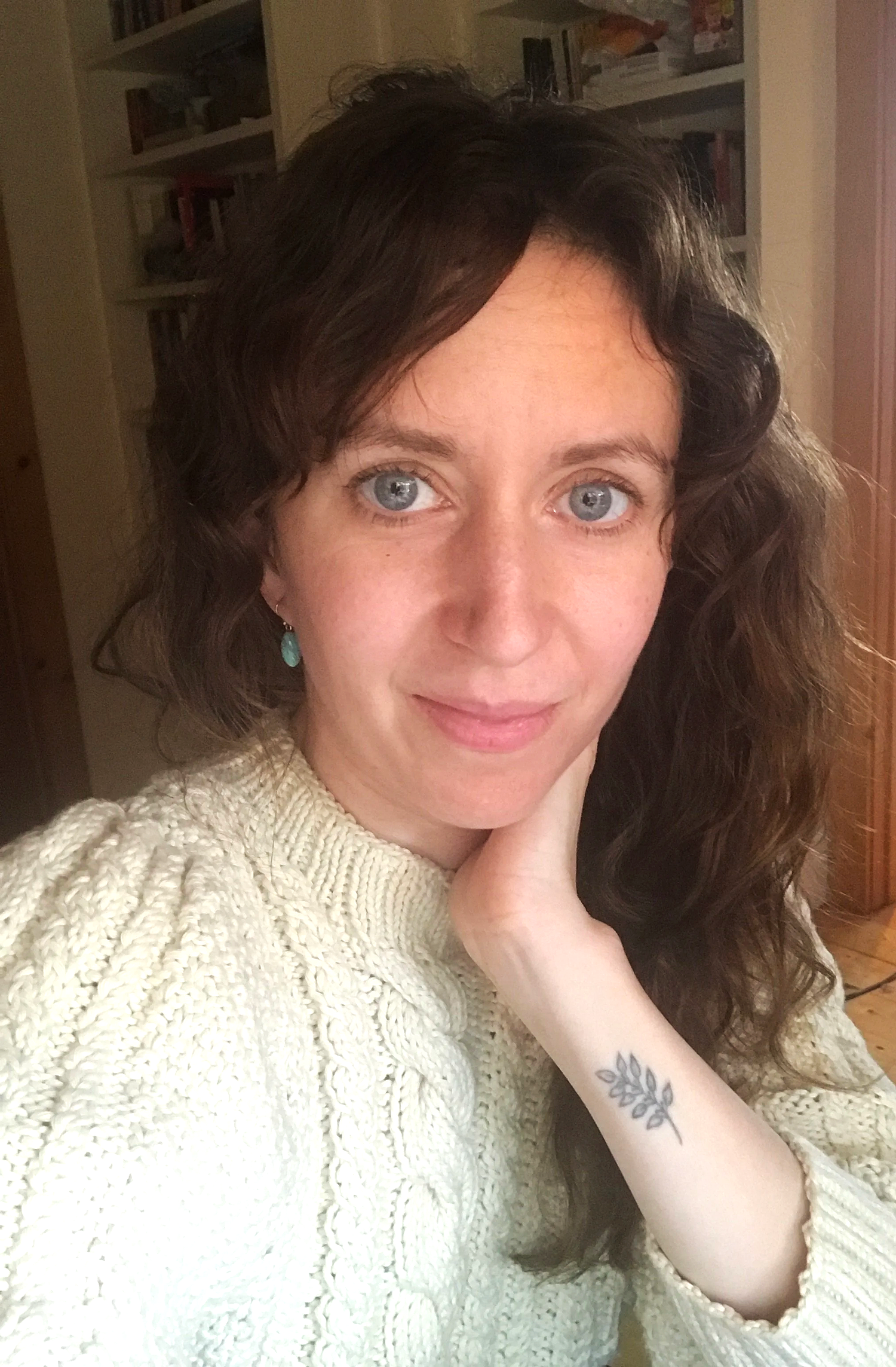 A woman with curly dark hair and light blue eyes, wearing a cream-colored knitted sweater and turquoise earrings, resting her head on her hand indoors.