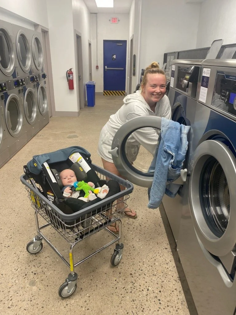 A woman smiling while washing clothes in a laundromat, with a baby in a car seat in a shopping cart nearby.