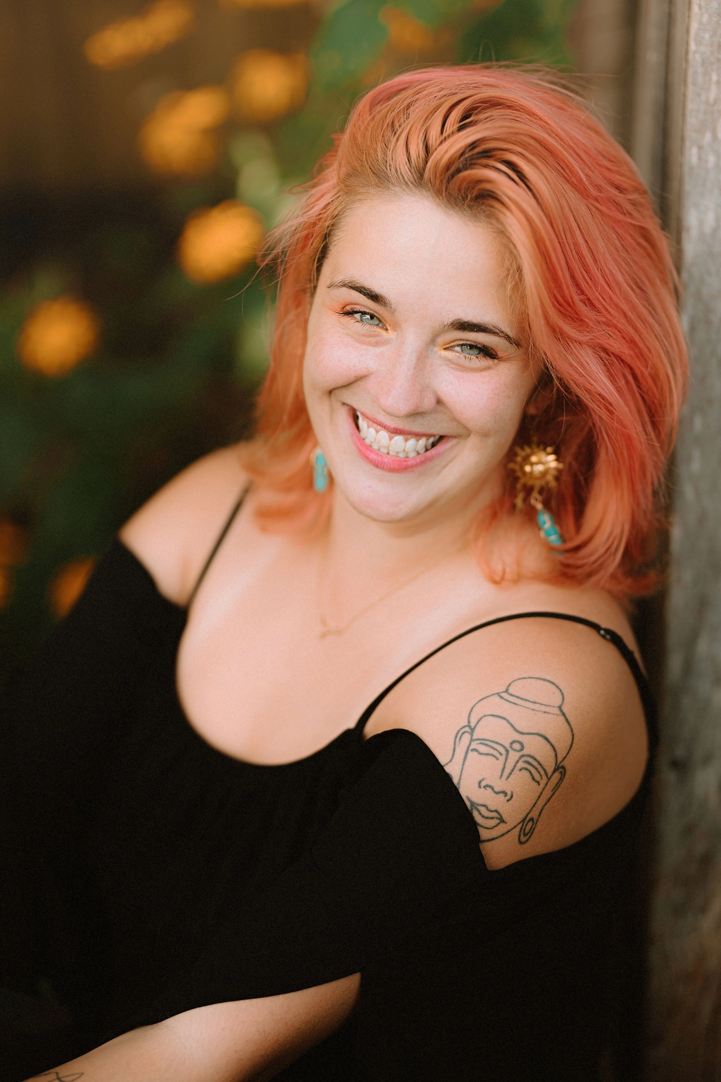 A woman with pink and orange hair smiling and wearing turquoise earrings, a black spaghetti strap top, and a gold necklace, leaning against a textured wall, with a tattoo of a serene face on her upper arm.