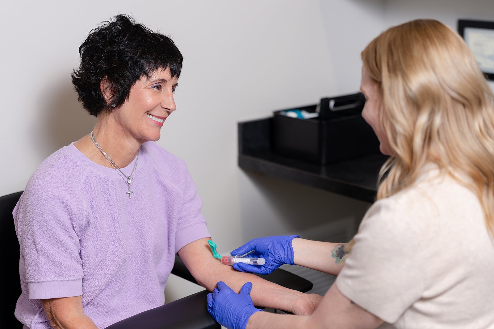 A woman in a purple shirt is getting her blood drawn by a healthcare professional wearing blue gloves in GevityMed office.