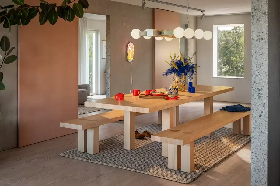 A modern dining room with a wooden table, two matching benches, and a rug underneath. The table has red mugs, a vase with blue and yellow flowers, and some plates. A contemporary light fixture with globe bulbs hangs above. There is a large window on 