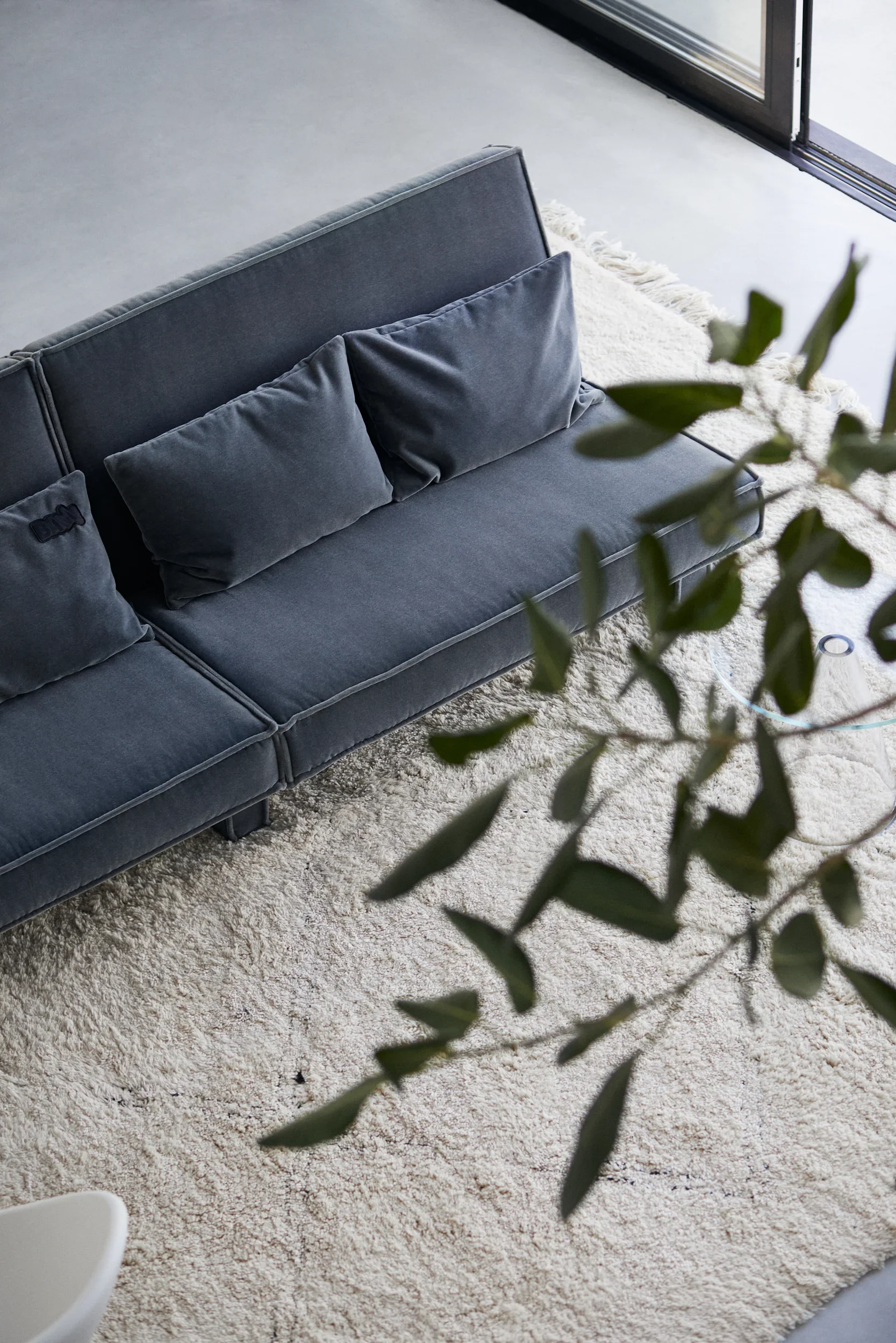 A modern living room with a grey velvet sofa on a white shag rug, near glass sliding doors, and some green plant leaves in the foreground.
