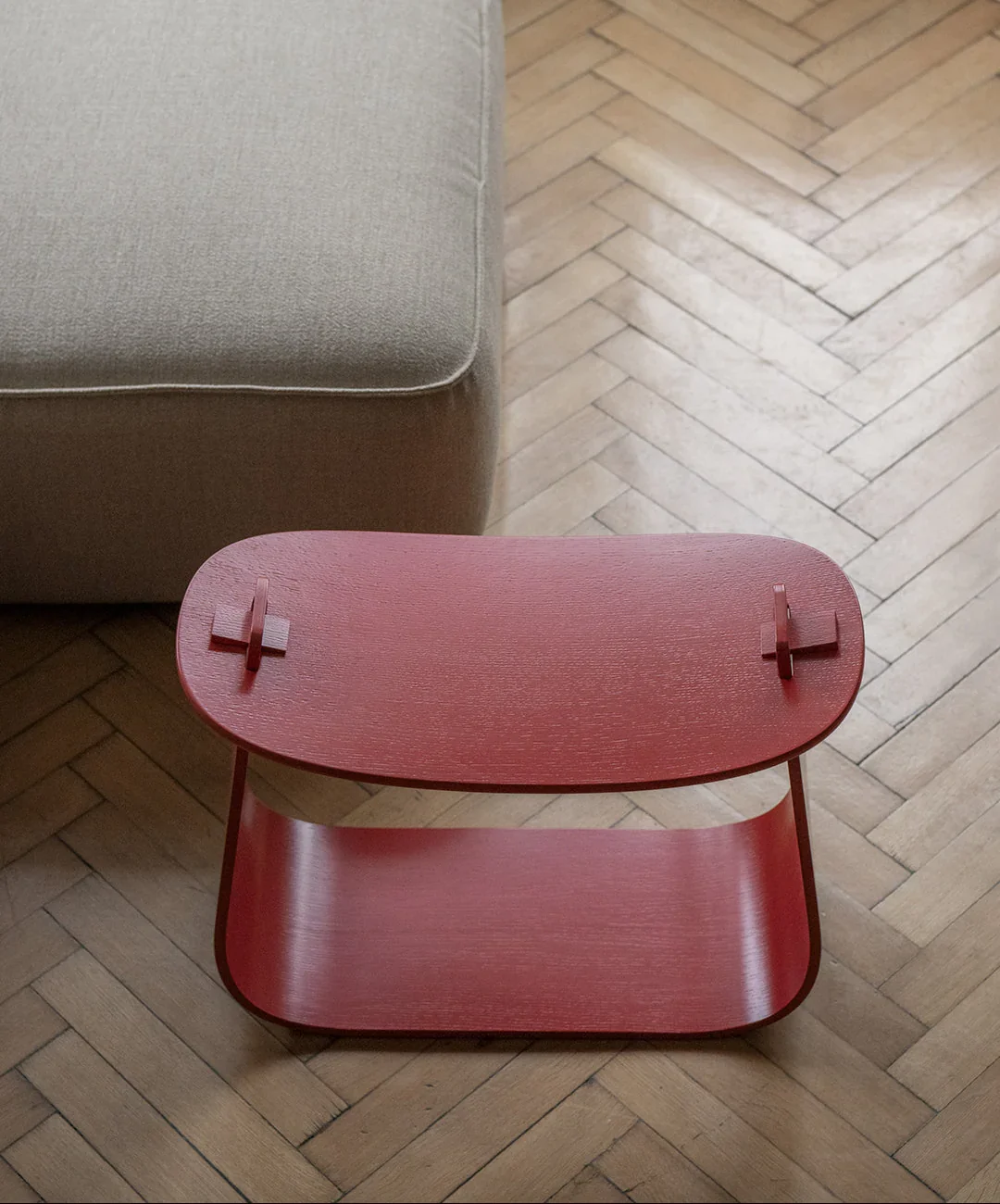 A red wooden side table with a curved top and two small raised handles, placed on a wooden herringbone floor next to a beige sofa.