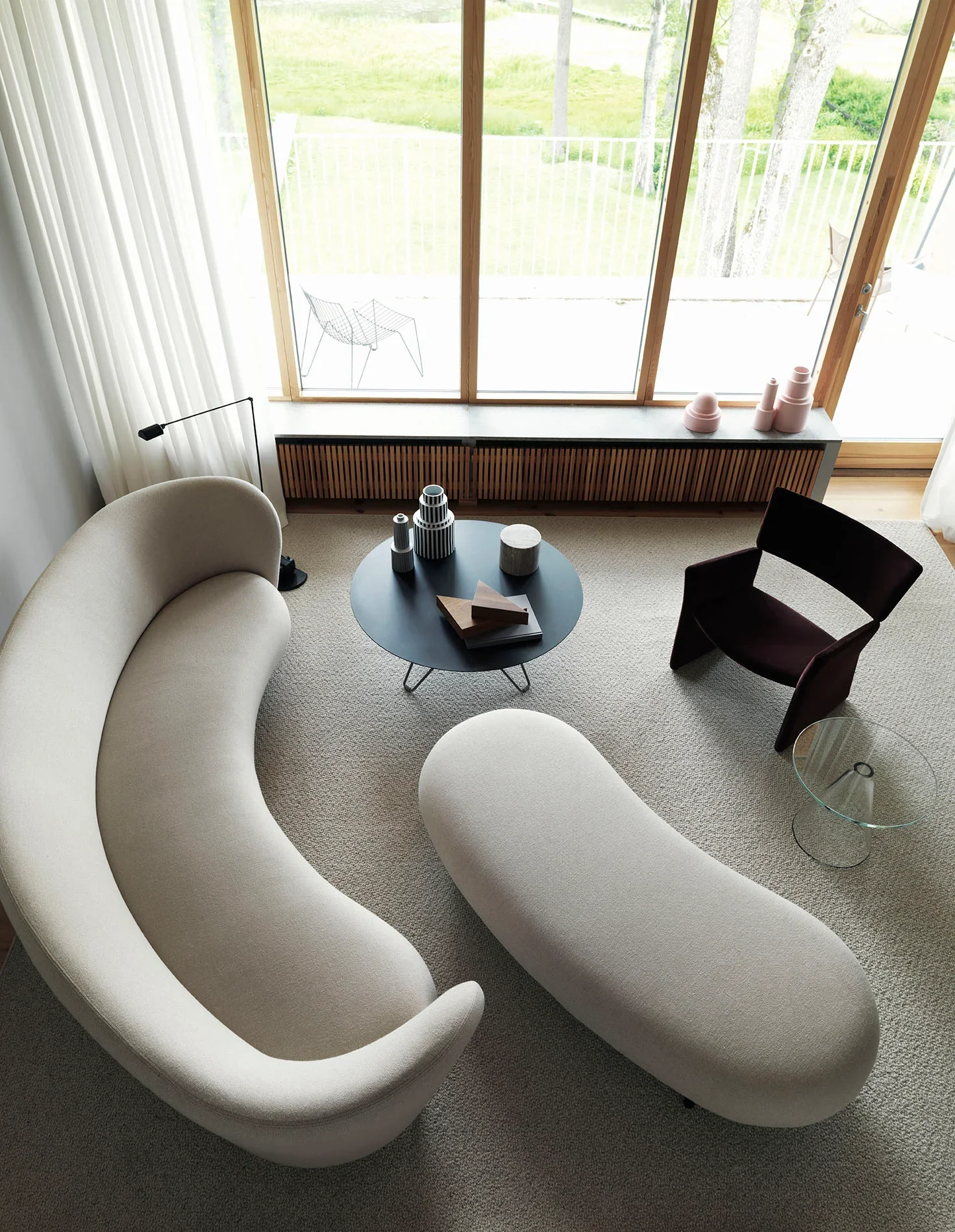 Living room with curved white sofa, black round coffee table with decor, side chair, small glass table, large window with view of outdoor patio and greenery, pink vases on window sill.