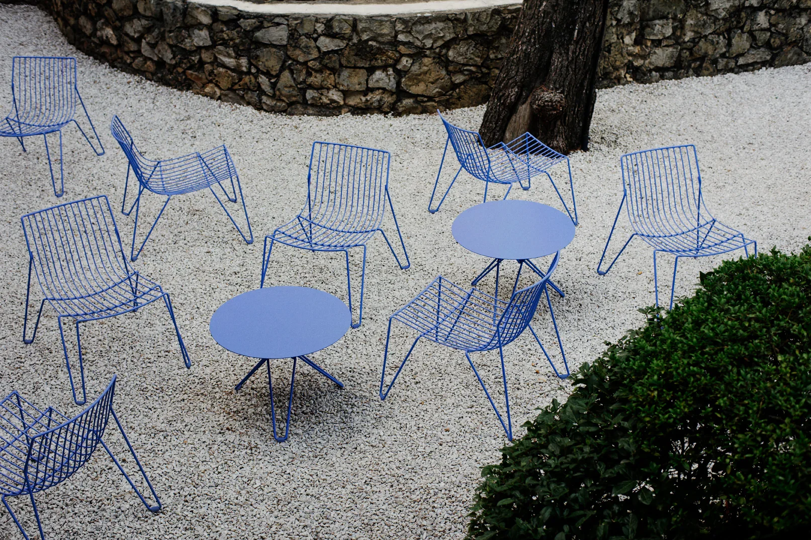 Outdoor seating area with blue wire chairs and round tables on white gravel, with a stone wall and tree in background and greenery in foreground.
