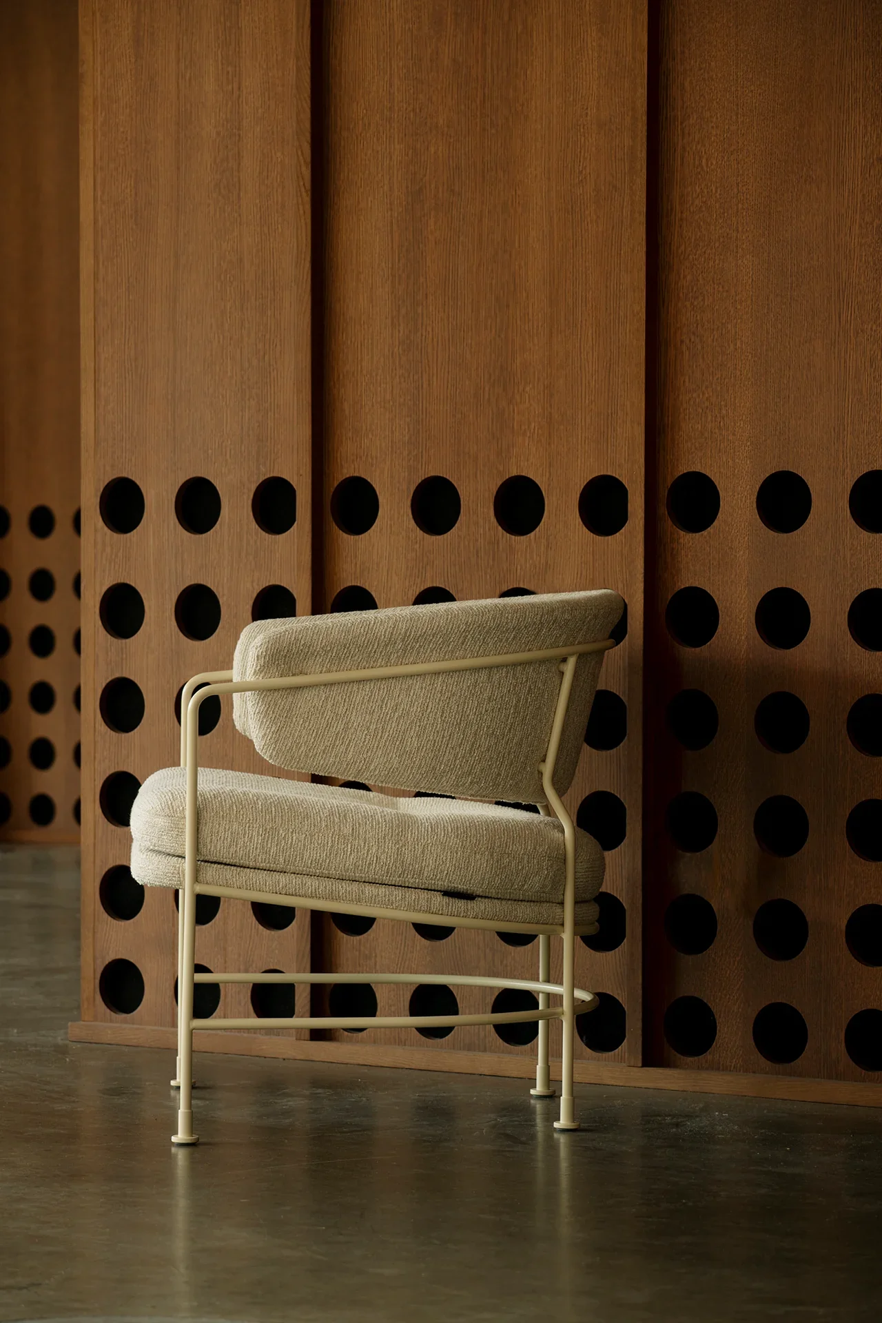A modern beige upholstered chair with a metal frame in front of a wooden room divider with circular cutouts.