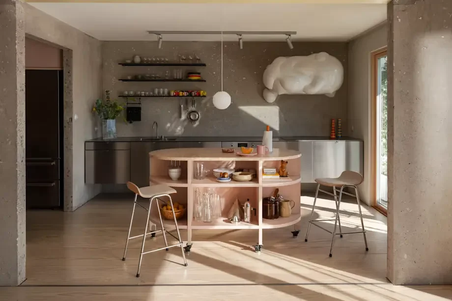 Modern kitchen with open layout, gray walls, minimalist shelves with dishes, a pink kitchen island on wheels, two bar stools, a large cloud-shaped light fixture, and a sliding glass door letting in natural light.