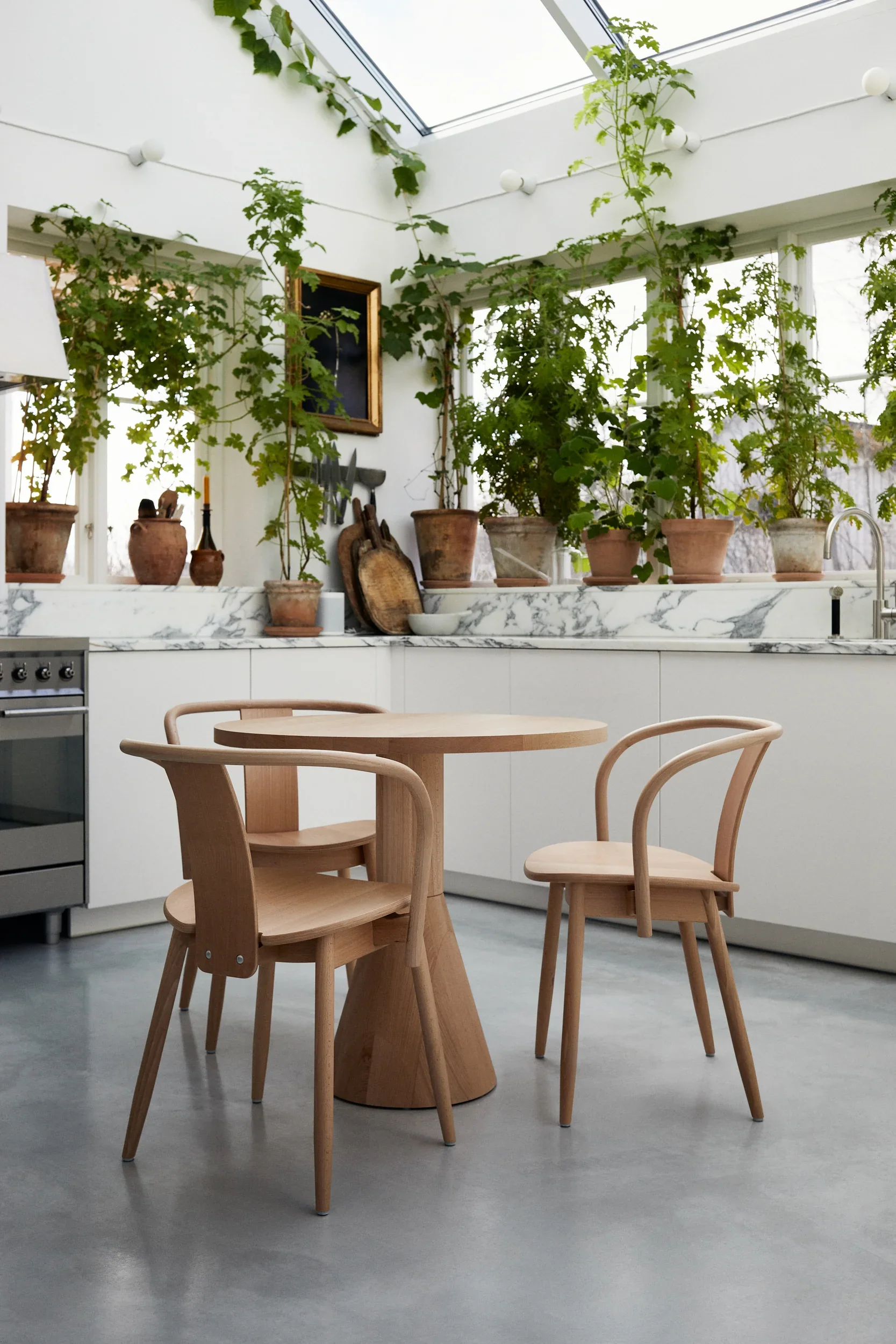 A modern kitchen with a marble countertop, potted plants on the windowsill, and wooden chairs around a small wooden table.
