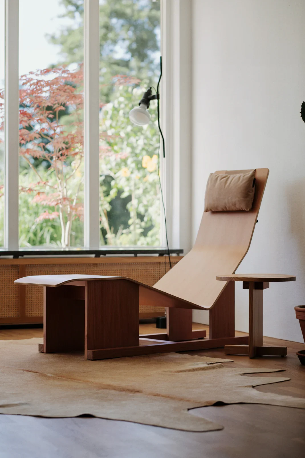 Interior of a room with a modern wooden chair and a small wooden side table, placed on a beige rug, near a window with greenery outside.