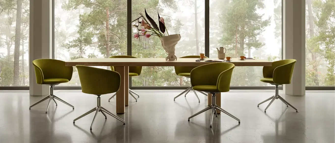 Modern dining room with a wooden table, six green swivel chairs, a vase with pink and dark flowers, and large floor-to-ceiling windows overlooking a wooded area.