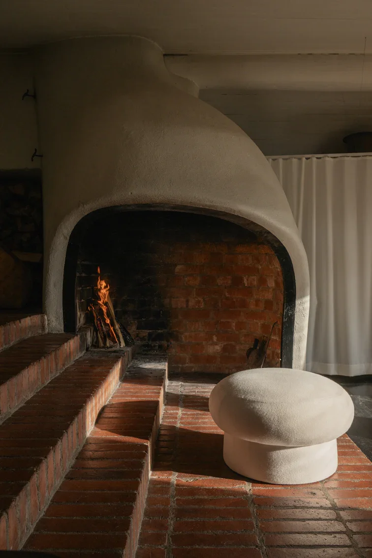A cozy indoor fireplace with a small fire burning inside, built with brick and concrete, accompanied by a rounded, white, textured stool on a brick floor.