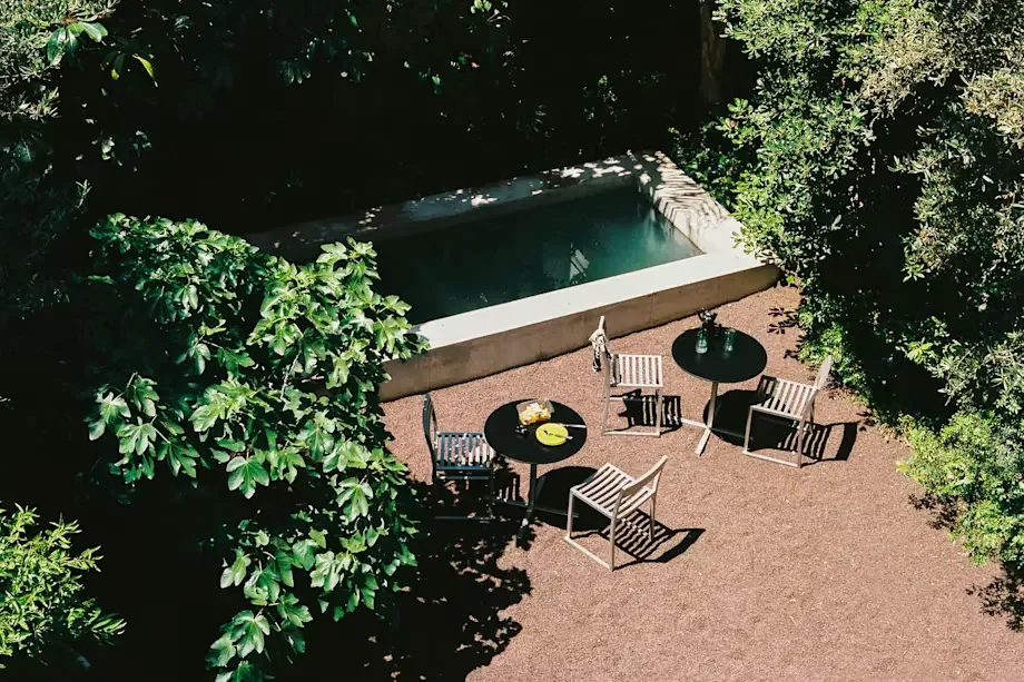 A backyard patio with two black round tables, each surrounded by chairs, and a small hot tub or pool in the background, partially shaded by trees.