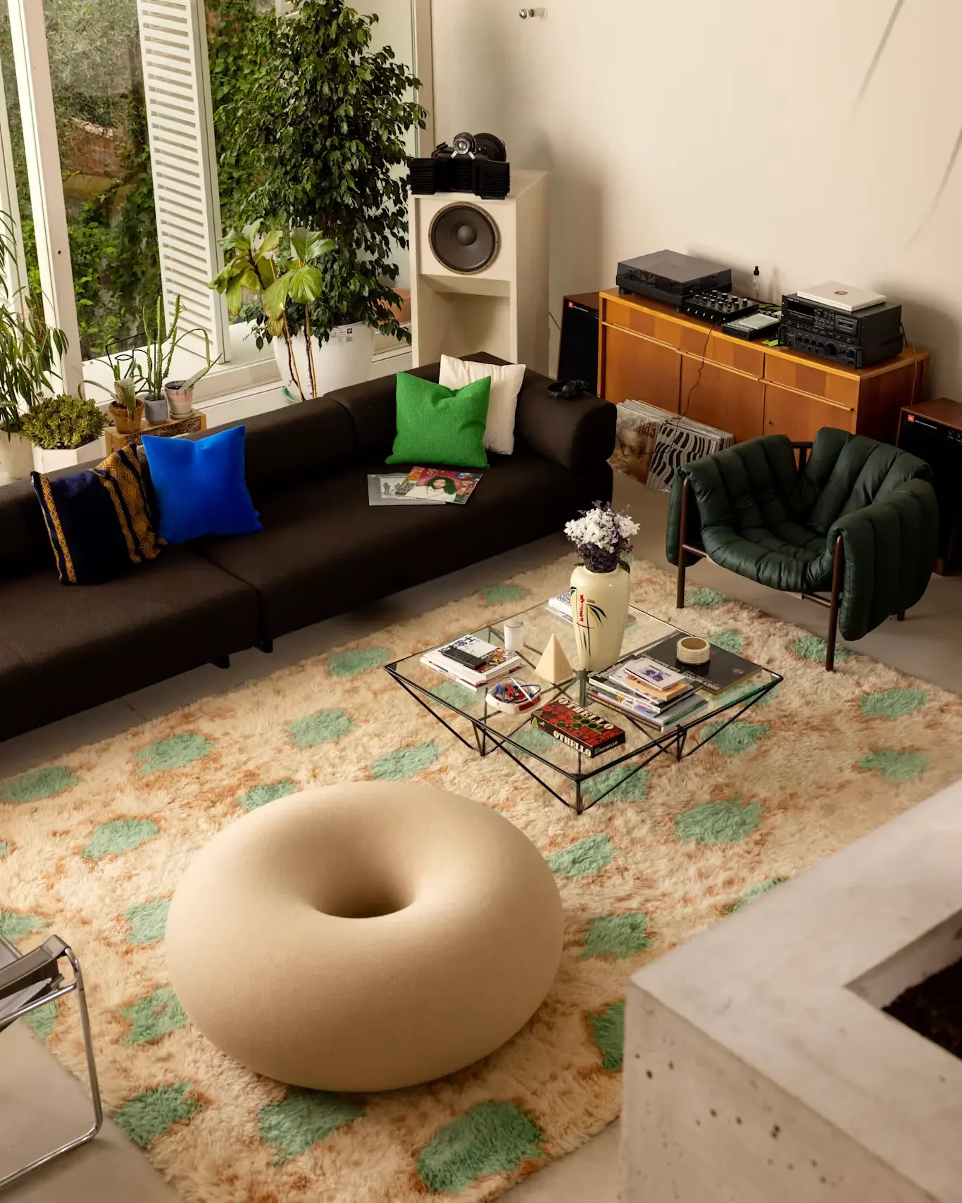 Living room with black sofa, green armchair, glass coffee table, plush carpet, potted plants, large window with open shutters, stereo equipment, and a vase with white flowers.
