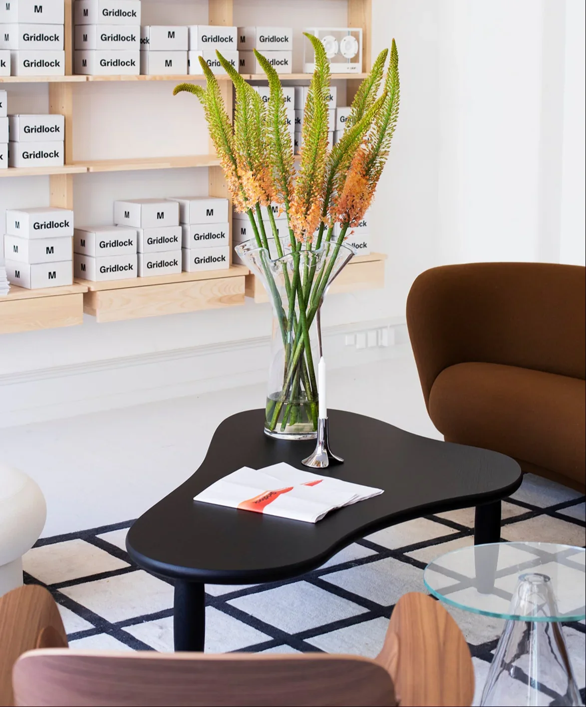 A living room with a black coffee table holding a glass vase with orange and green flowers, a white candle, and a cloth napkin. To the right, part of a brown armchair and a glass side table are visible. Background features a wall with wooden shelves 