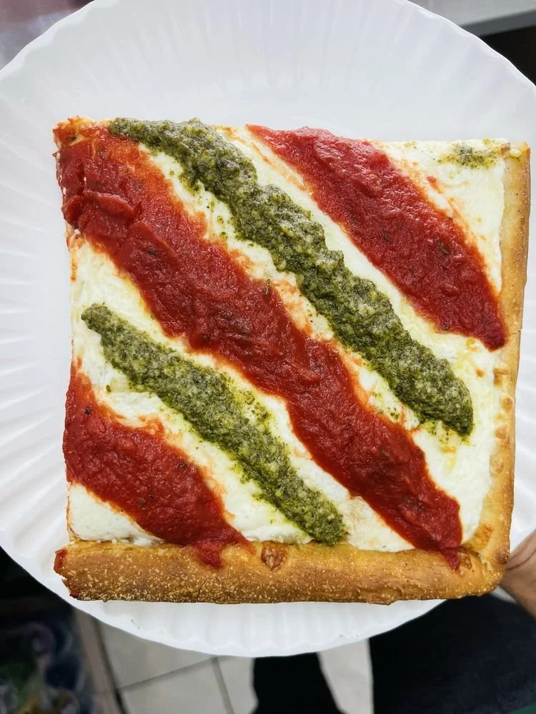 Slice of pizza with a thick crust, topped with tomato sauce, cheese, and green pesto sauce in a striped pattern.