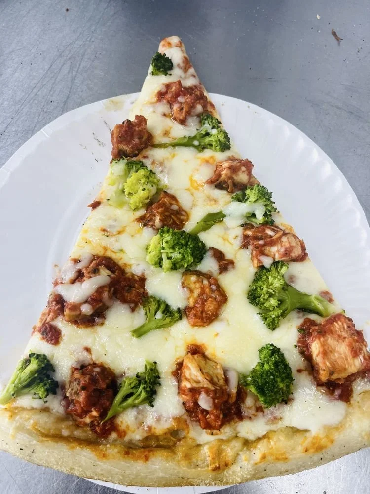 Close-up of a slice of pizza topped with broccoli, chicken, and melted cheese on a white plate.