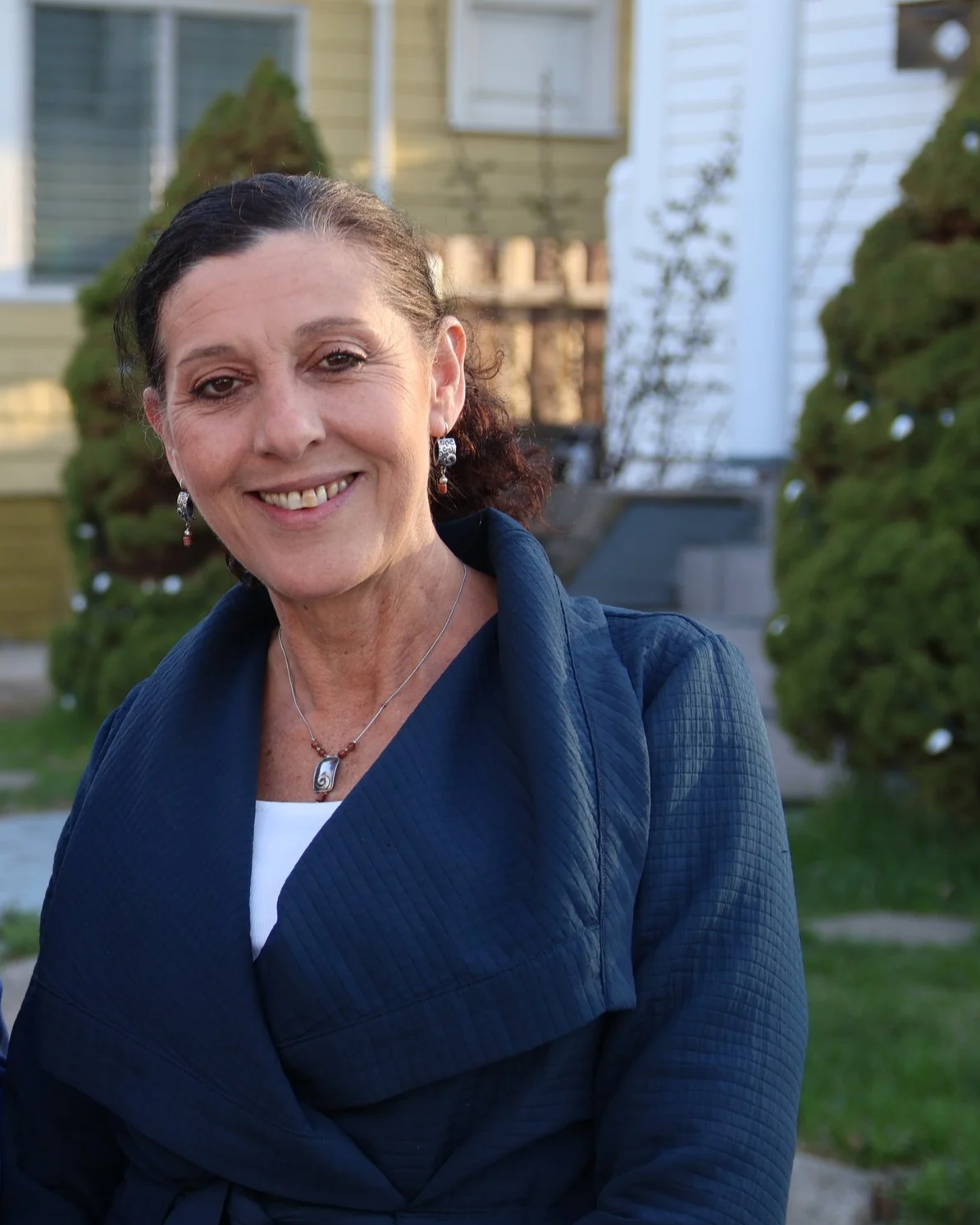 Amy, a  smiling woman with dark curly hair, wearing a navy blue jacket and jewelry, standing outdoors in front of a house with trees and greenery.
