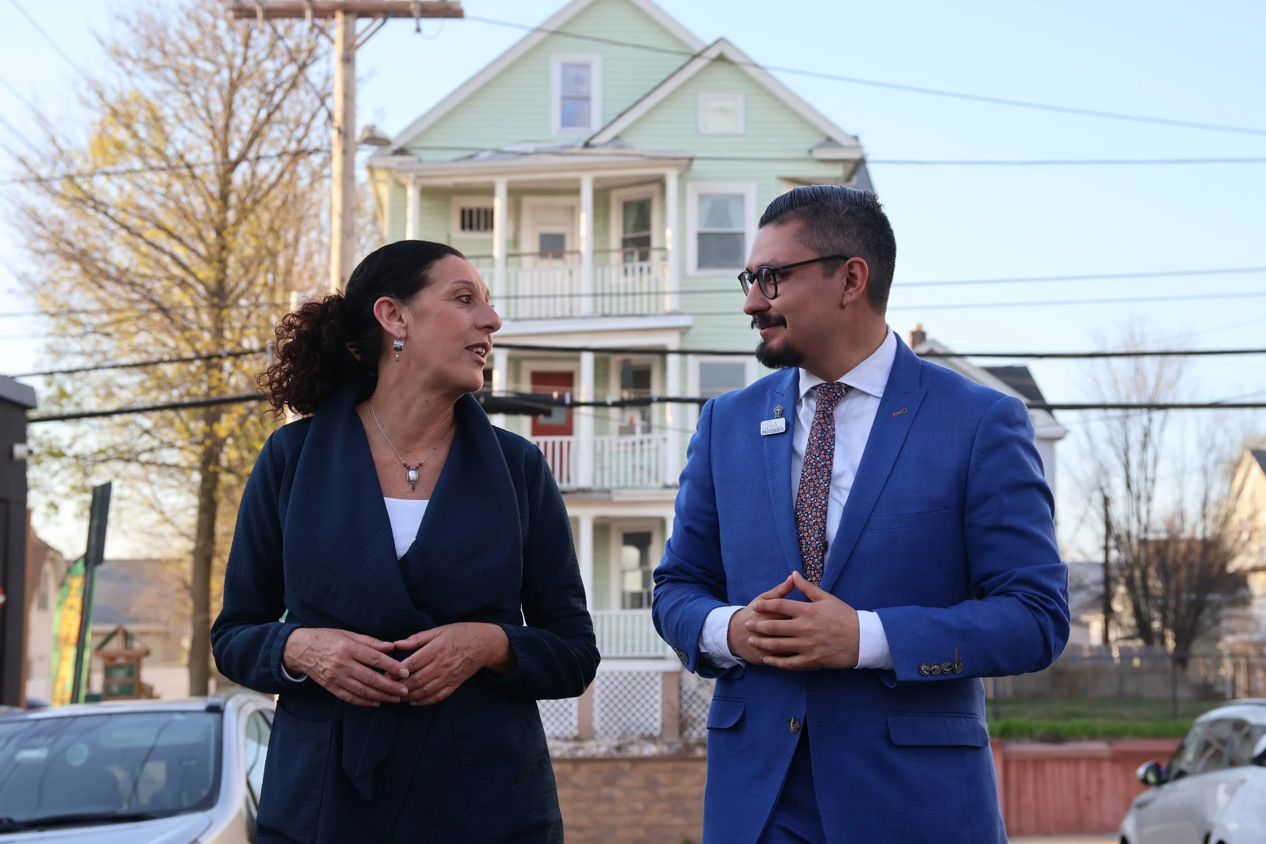 Two people, a woman and a man, are standing outside in front of a green house in conversation. The woman has dark, curly hair and is wearing a dark blazer, while the man has short dark hair, glasses, and is dressed in a bright blue suit with a patterned tie. They are looking at each other and smiling. There are cars parked behind them and trees with autumn-colored leaves.