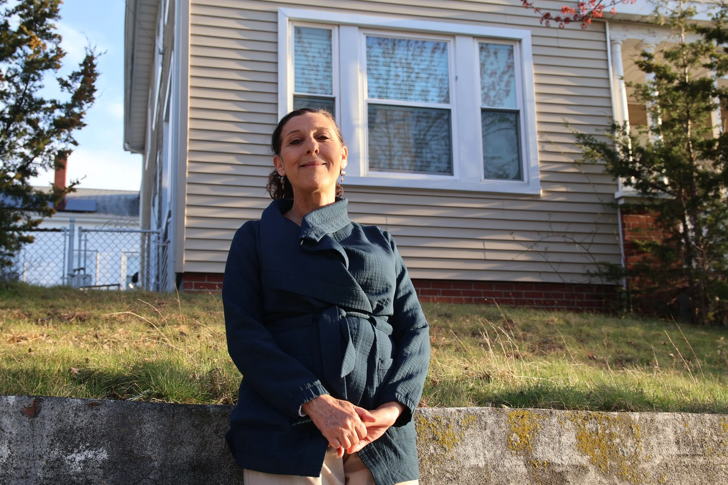 A woman sitting outdoors on a low stone wall in front of a house with beige siding, multiple windows, surrounded by trees and greenery, during daytime.