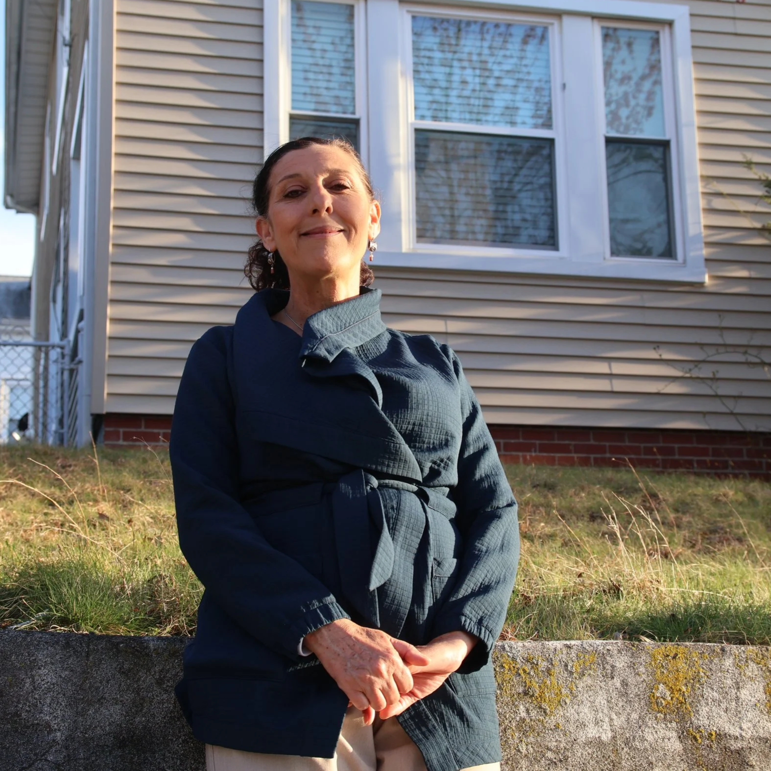 A woman sitting outdoors in front of a house with beige siding and large white-framed windows. She is smiling, wearing a dark jacket, earrings, and has her hands clasped in her lap. The grass and a low concrete wall are visible in the foreground.