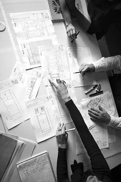 Plusieurs personnes en train de discuter autour d'une table avec des plans d'architecture, des croquis et des documents liés à la construction ou à l'architecture.