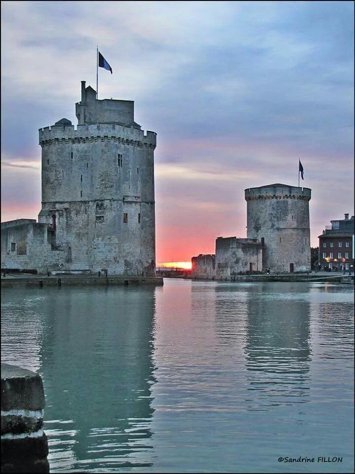 Deux tours médiévales entourées d'eau, avec un coucher de soleil en arrière-plan, sur un plan d'eau calme.
