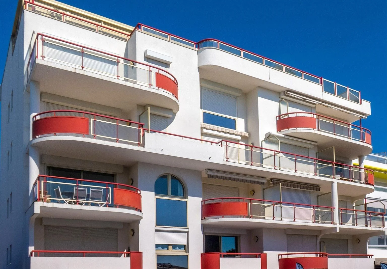 Immeuble blanc avec plusieurs balcons arrondis rouges, fenêtres avec volets roulants et un ciel bleu en arrière-plan.