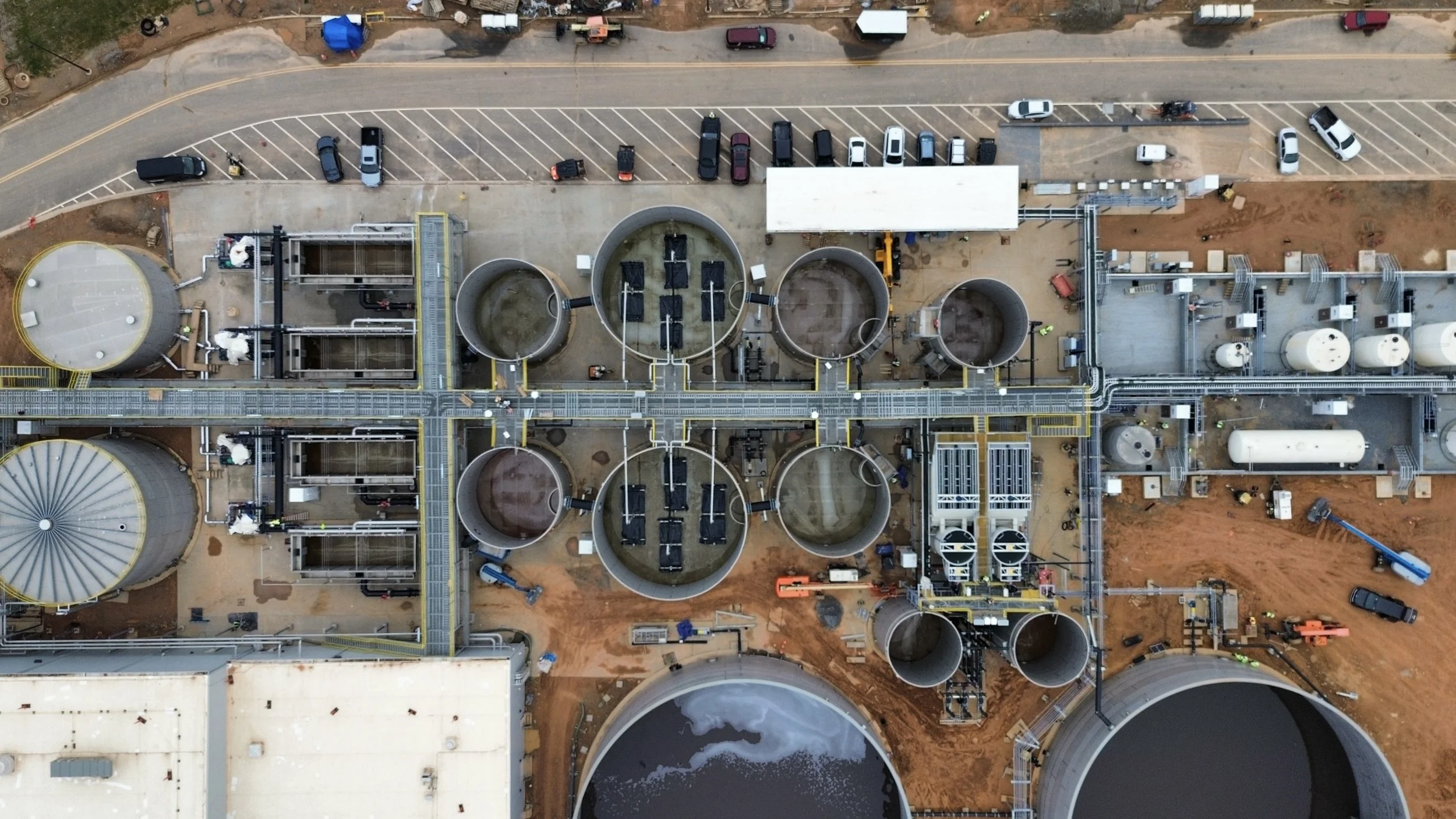 An aerial view of an industrial wastewater treatment plant with large circular and rectangular tanks, walkways, connected pipes, and parked vehicles.