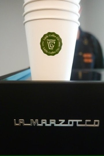 White disposable coffee cup with a green logo, placed on a black surface with 'La Marzocco' branding, against a blurred background.