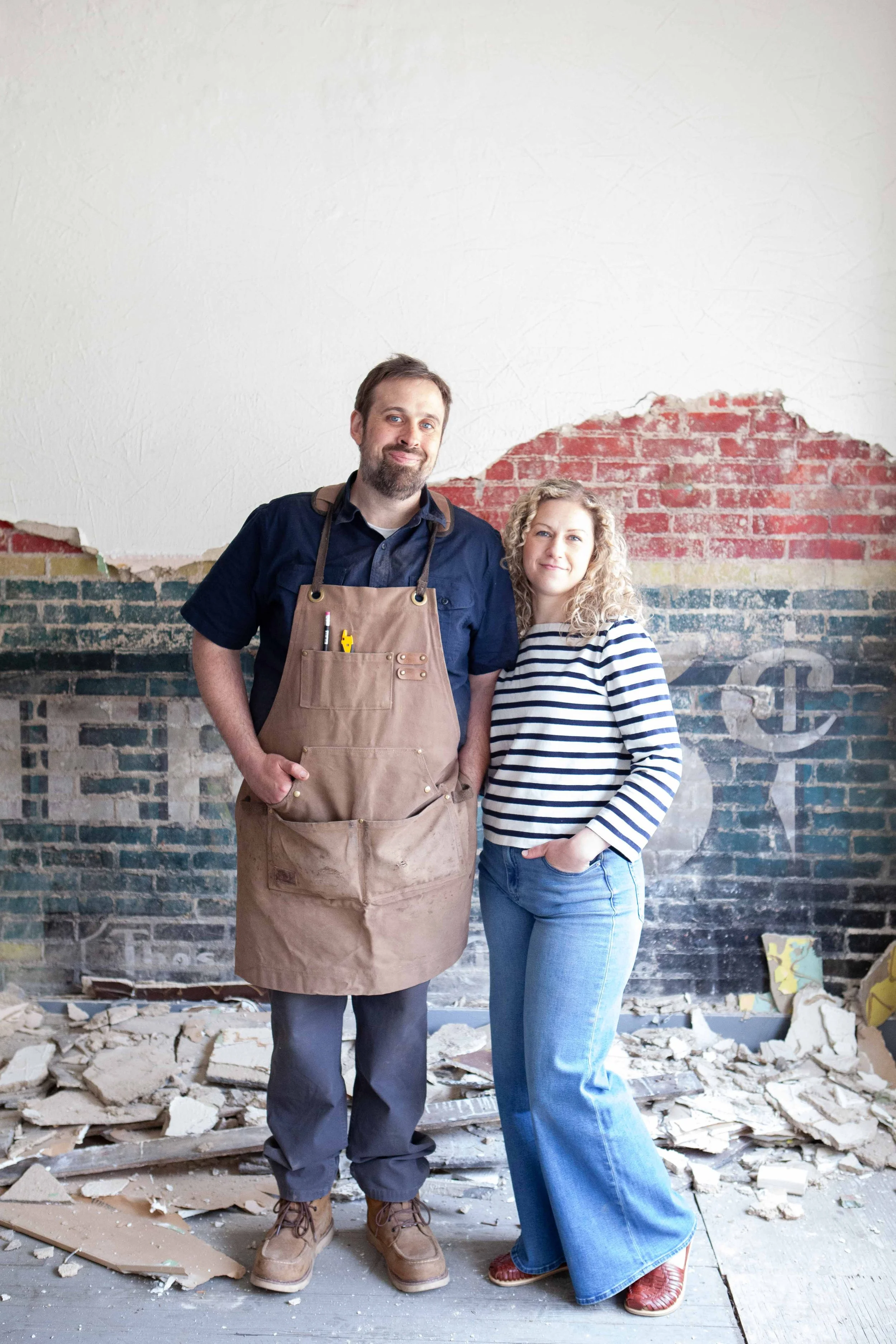 Man and woman standing together in front of a partially demolished wall with rubble on the floor, both wearing casual clothes and smiling.